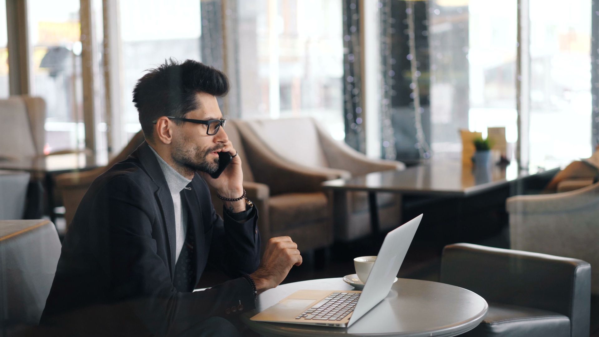 Business professional using smartphone and laptop in a café for a remote work meeting.