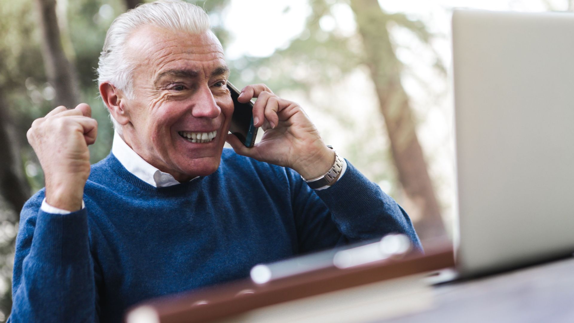 Elderly man smiling and cheering while talking on the phone using laptop outdoors.