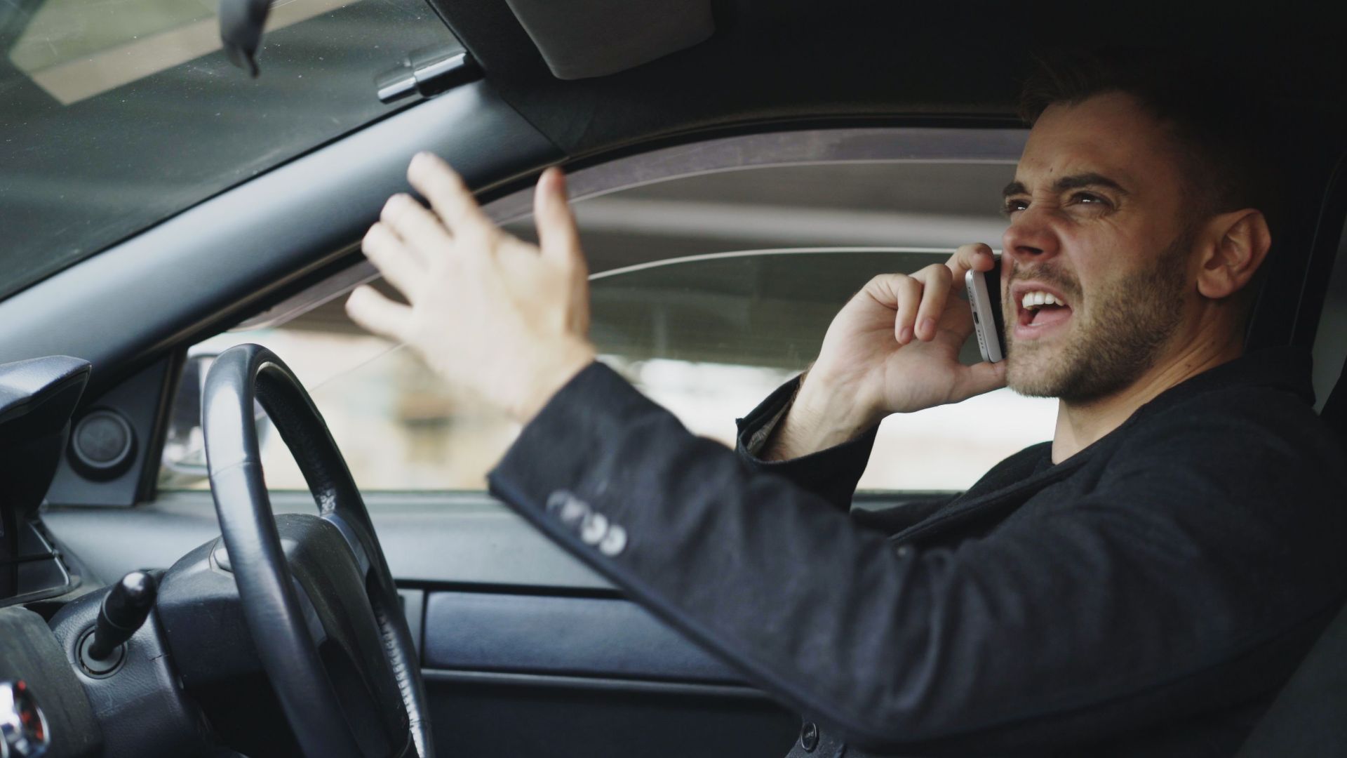 Stressed man gesturing while on phone in car, expressing frustration.