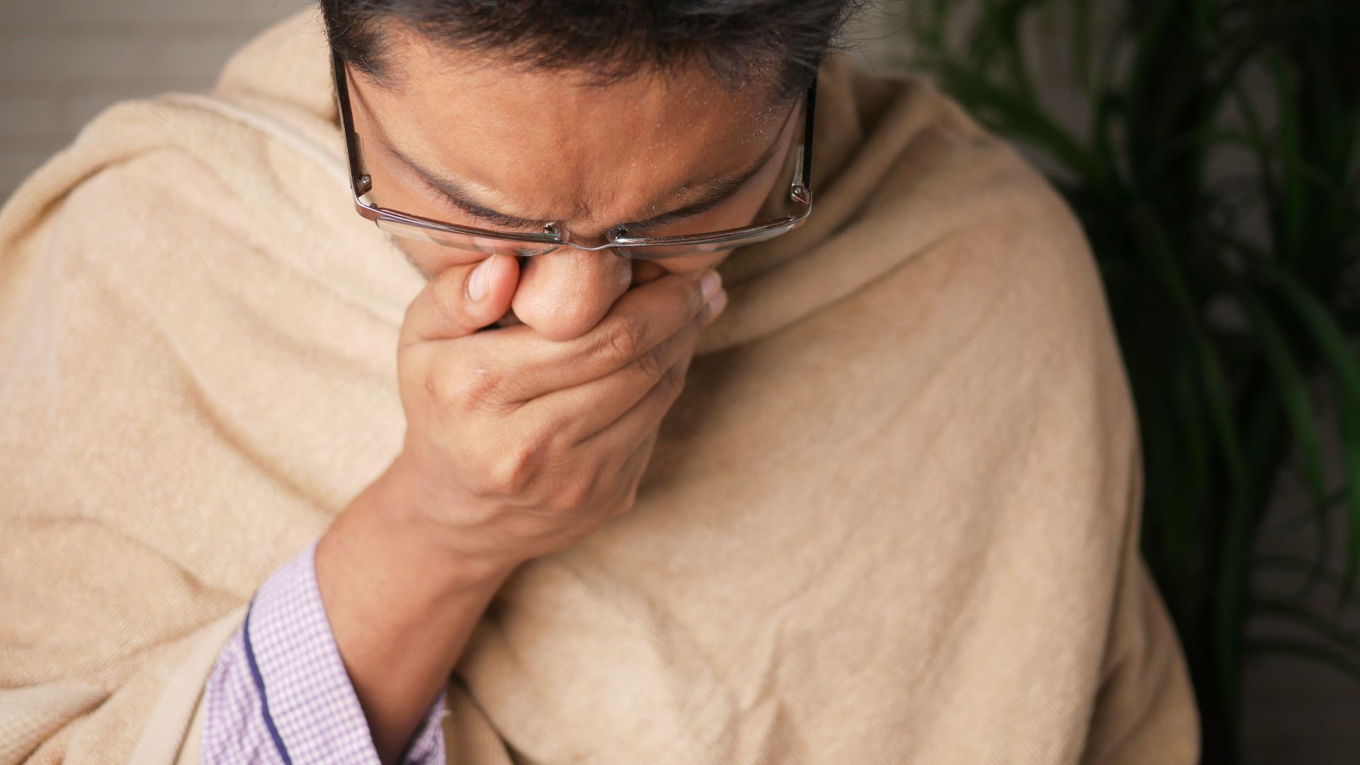 Adult wearing glasses coughs into hand indoors, indicating illness or flu symptoms.