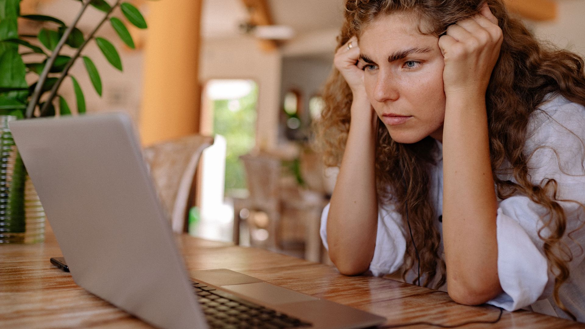 A woman with curly hair intensely focusing on her laptop indoors with plants and natural lighting.