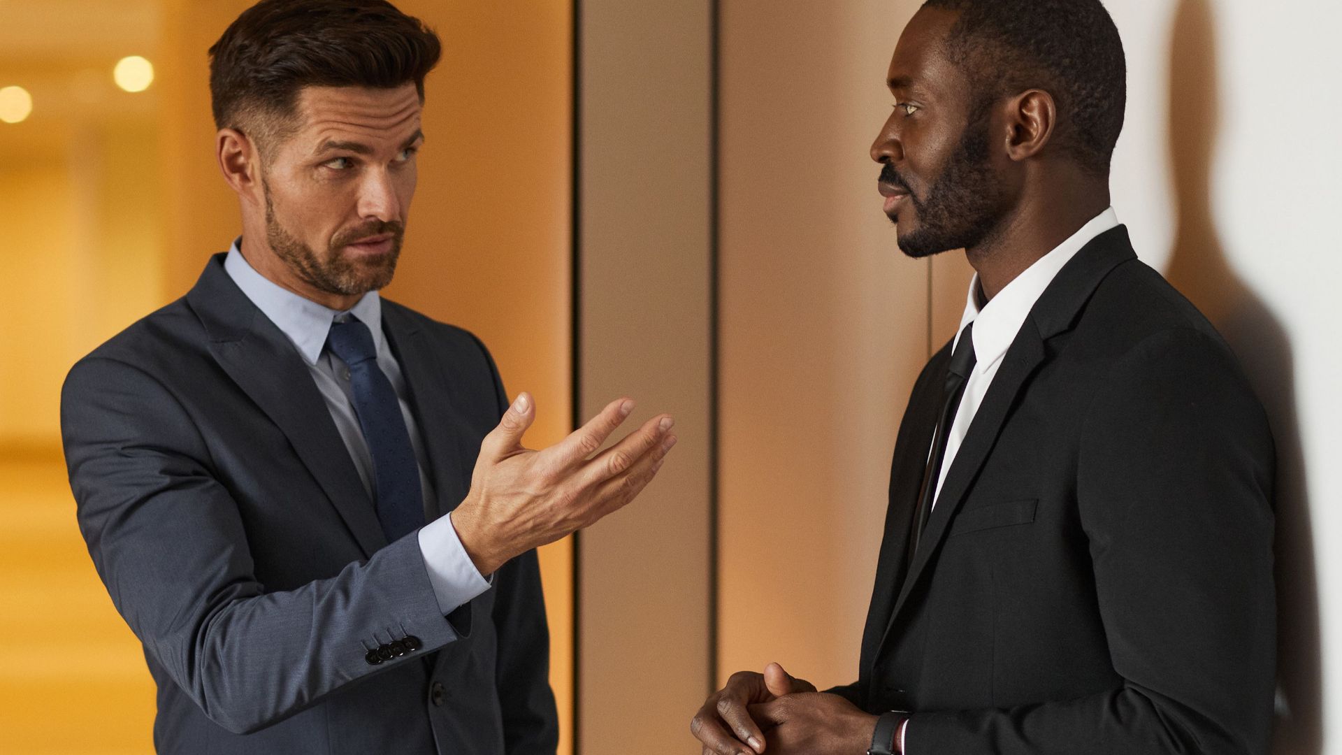 Two businessmen in formal attire engaged in a serious discussion in a modern office hallway.