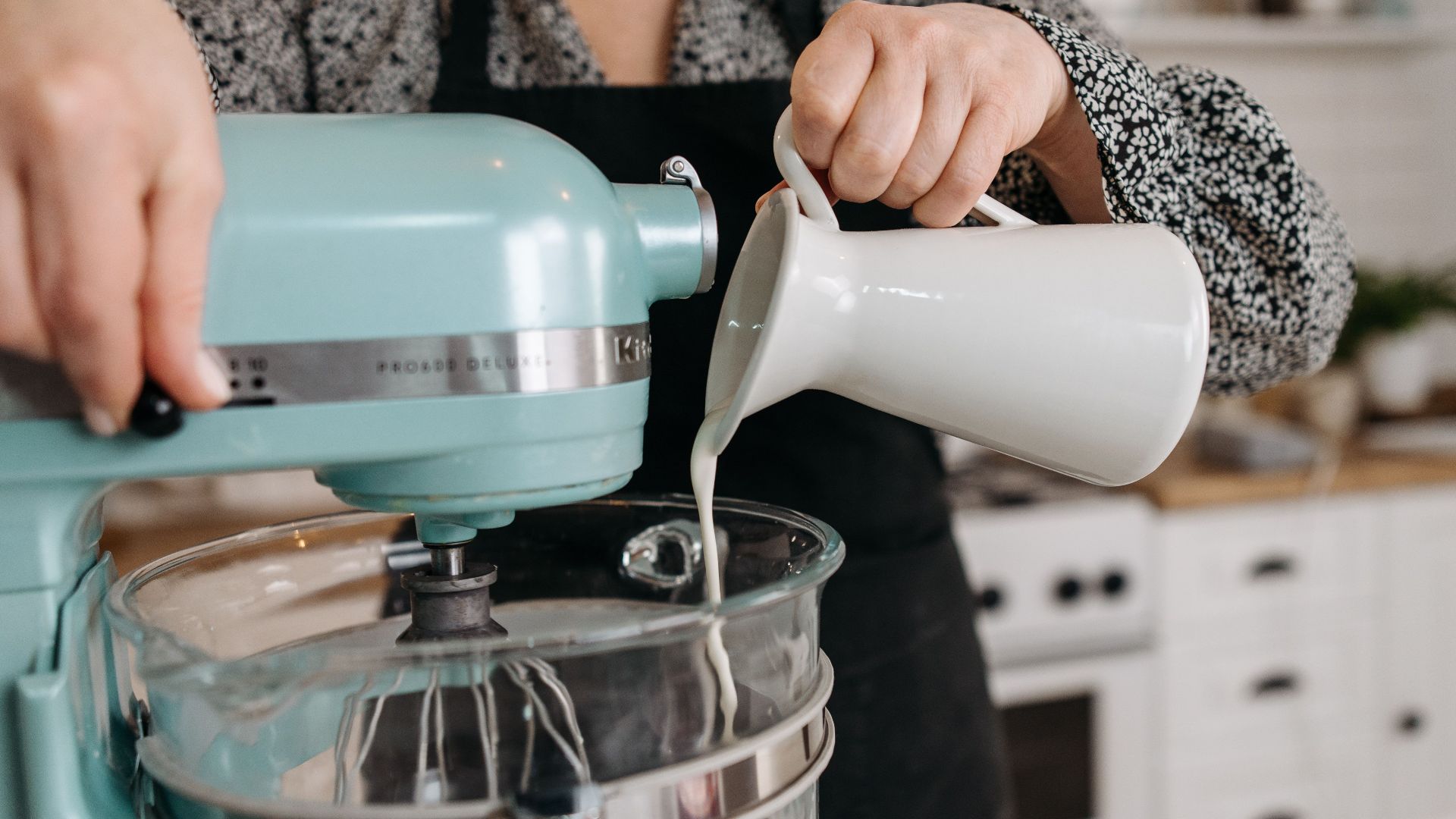 Person in kitchen pouring milk into a stand mixer bowl during baking preparation.
