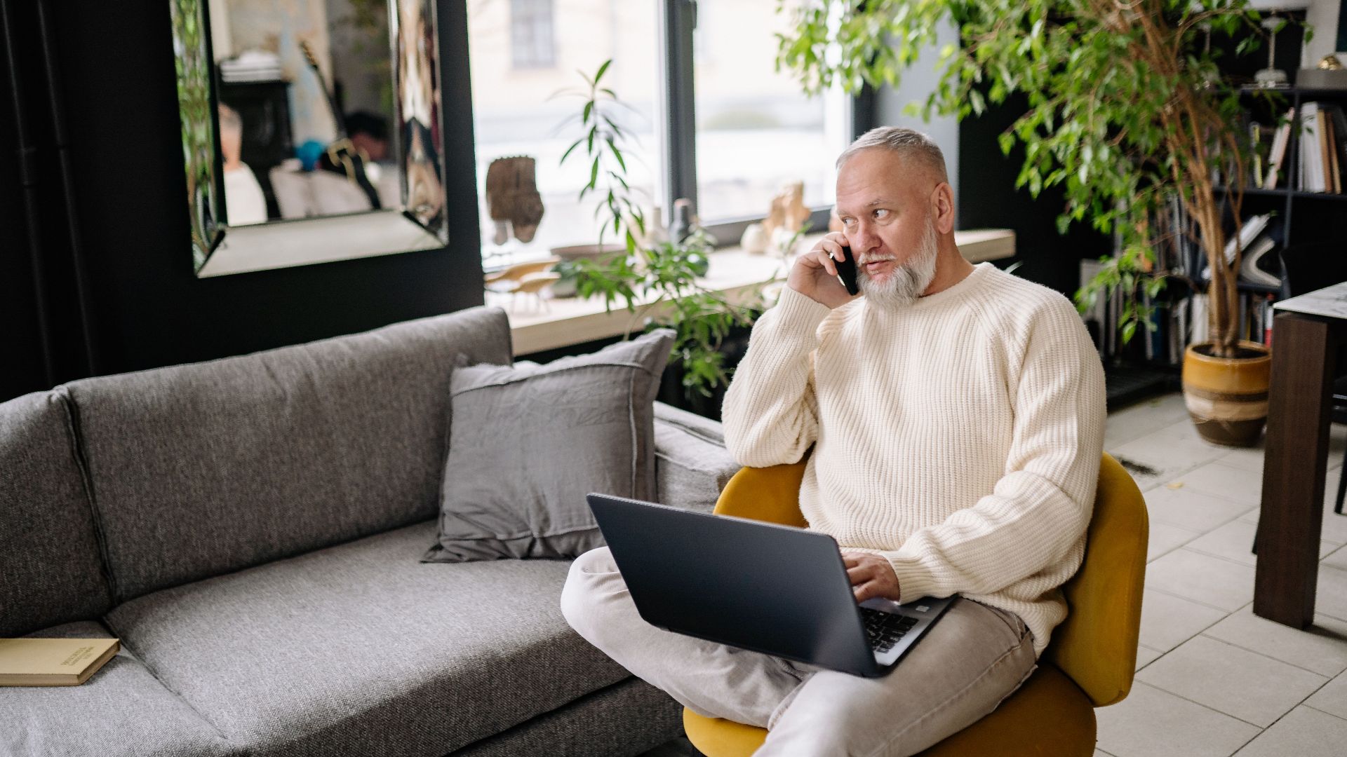 Elderly man multitasking at home, using laptop and phone, sitting comfortably indoors.