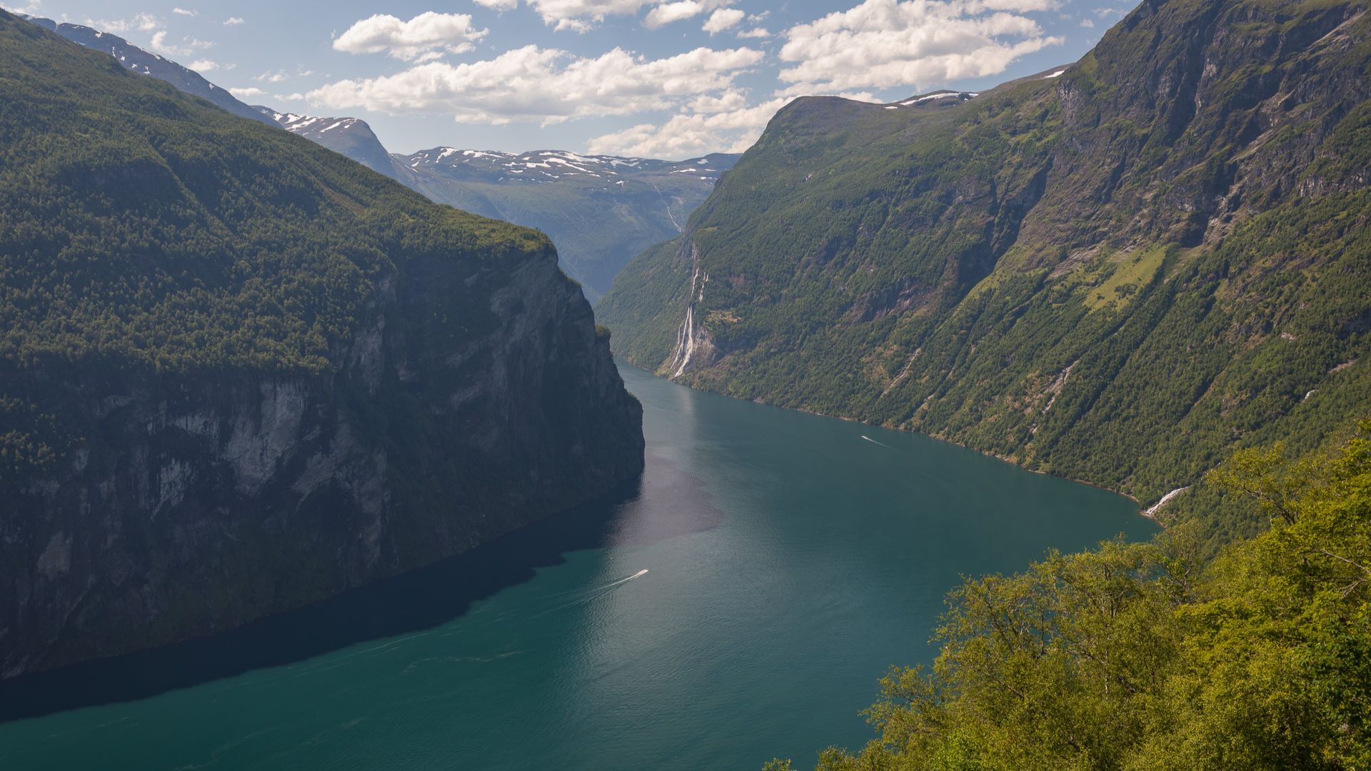Panoramic landscape view of Geirangerfjorden (Geiranger Fjord) in Stranda Municipality, Møre og Romsdal, Norway. The fjord cuts through dramatic, steep-sided mountains with patches of lingering snow on the high ridges. A tall waterfall can be seen cascadi