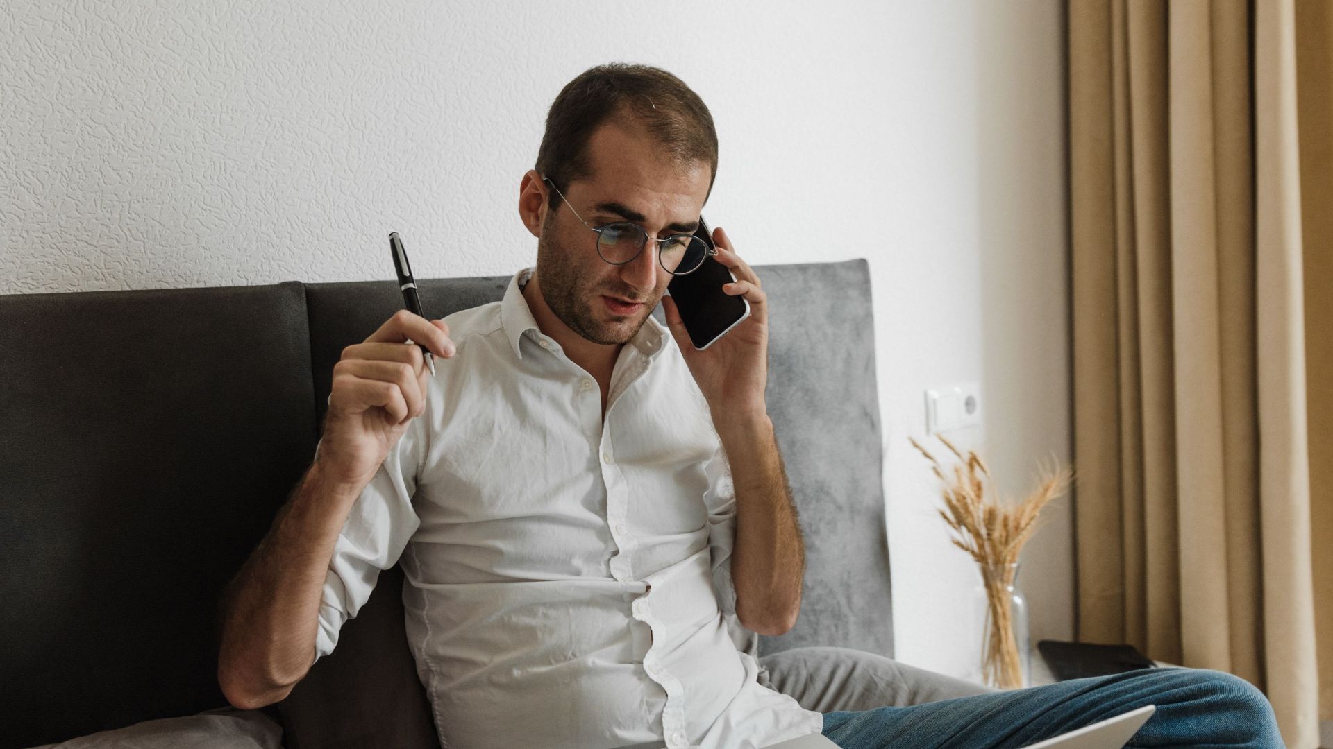 Adult male multitasking at home office, using laptop and smartphone while sitting on bed.