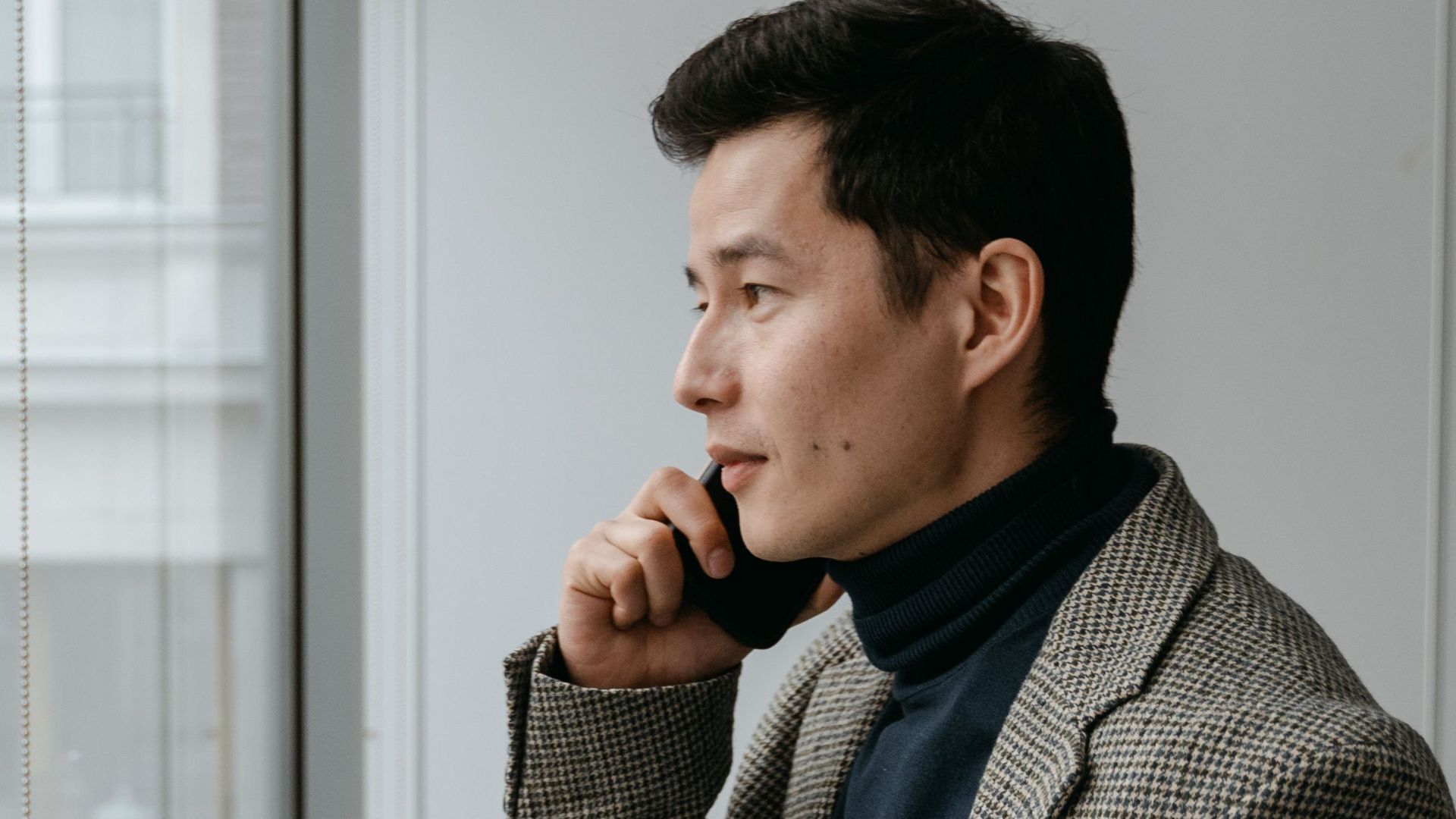 Confident businessman in a gray blazer talking on phone by office window.