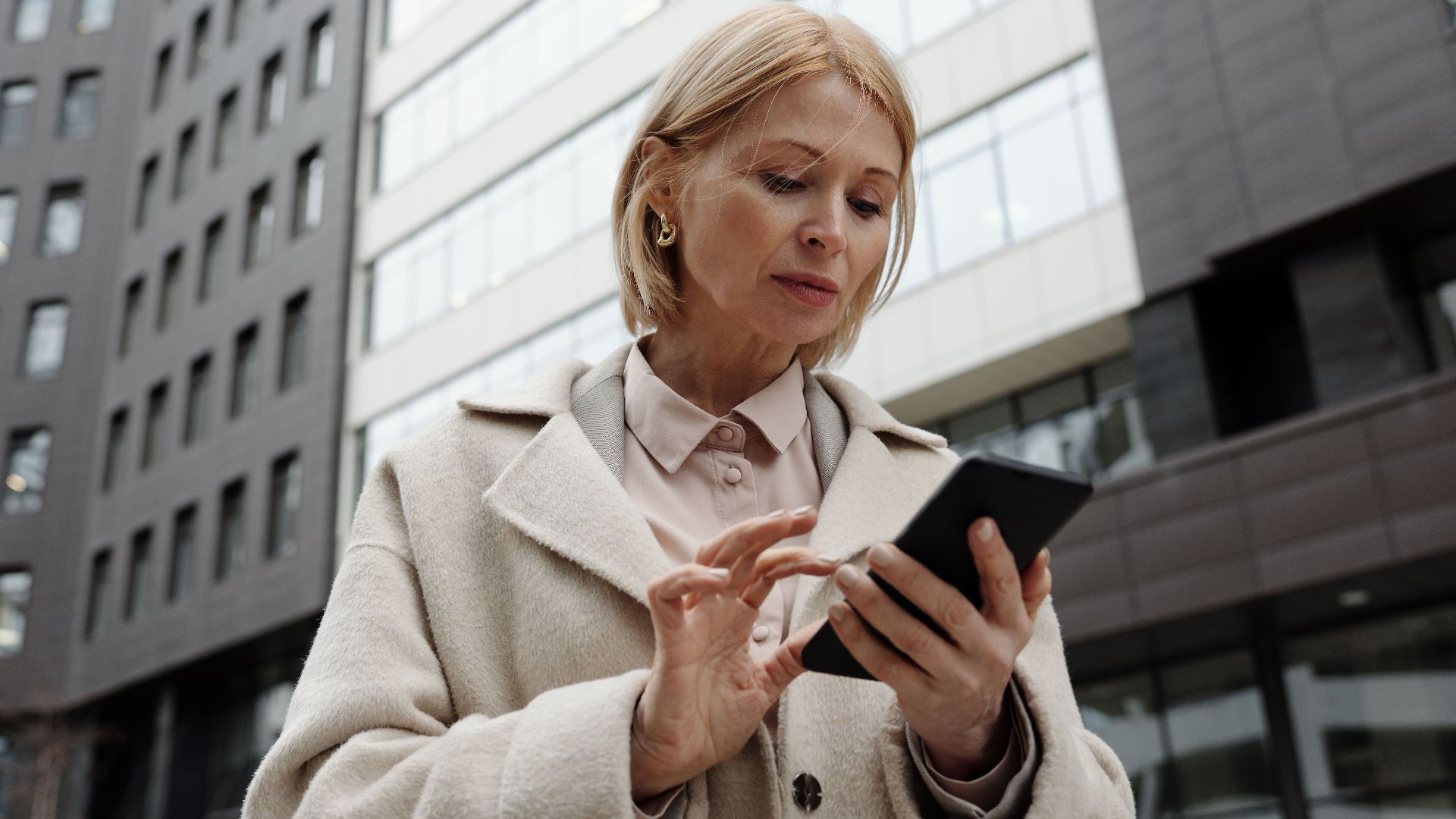 Woman in coat using smartphone in front of modern building. Professional and focused expression.