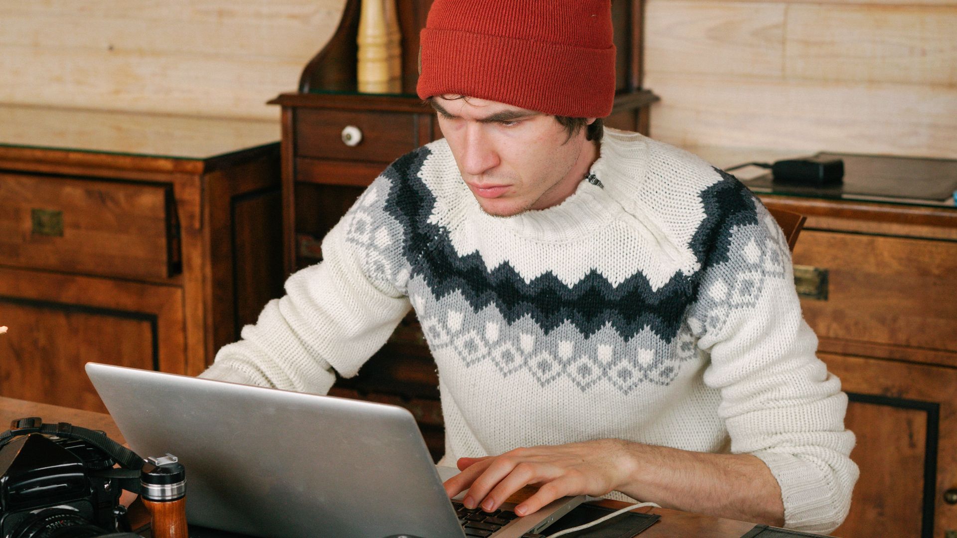 Man wearing sweater and beanie works on laptop at wooden table with cameras, in a cozy indoor setting.