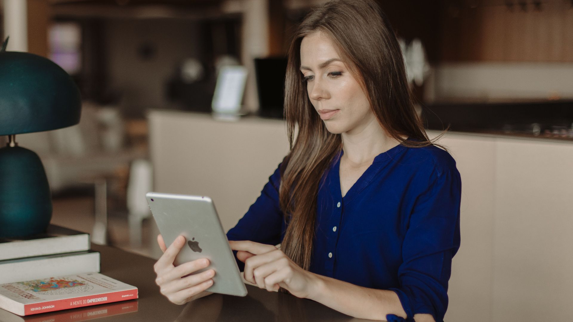 Woman reading on a tablet at a desk with books in a modern office setting.