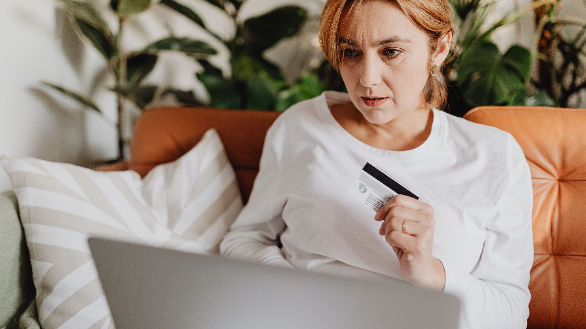A woman sitting on a couch shopping online with a credit card and laptop in a cozy living room.