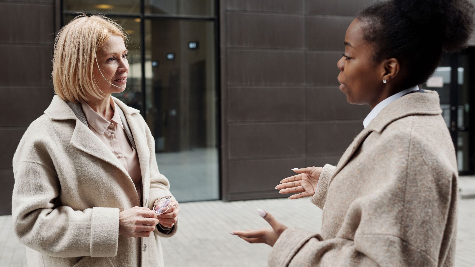 Two businesswomen in coats converse outside a modern office building.