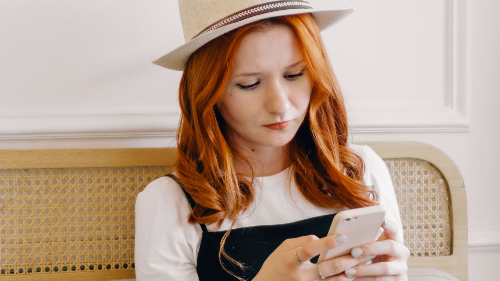 A young woman in a fedora hat texting on her smartphone while sitting with a laptop indoors.