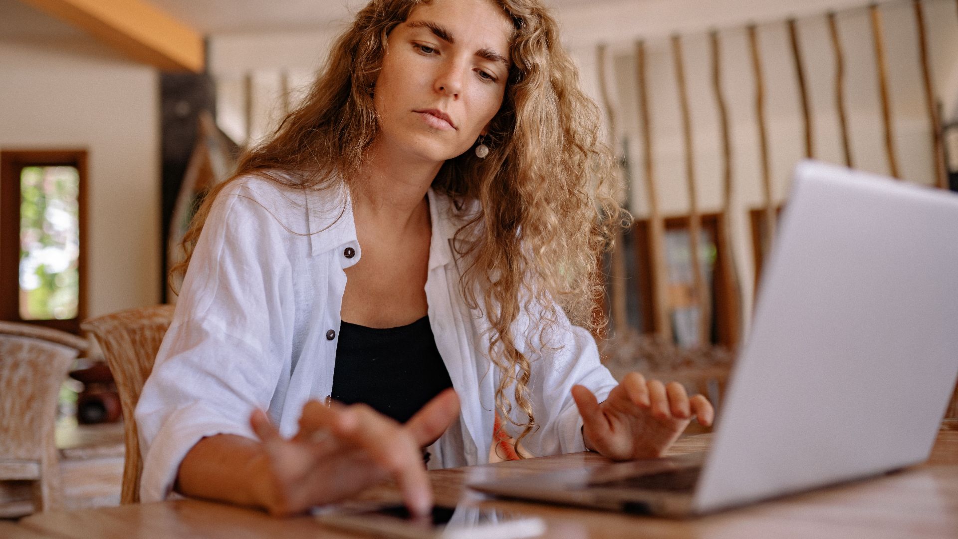 A woman intently works on a laptop while using a smartphone indoors, symbolizing modern remote work.