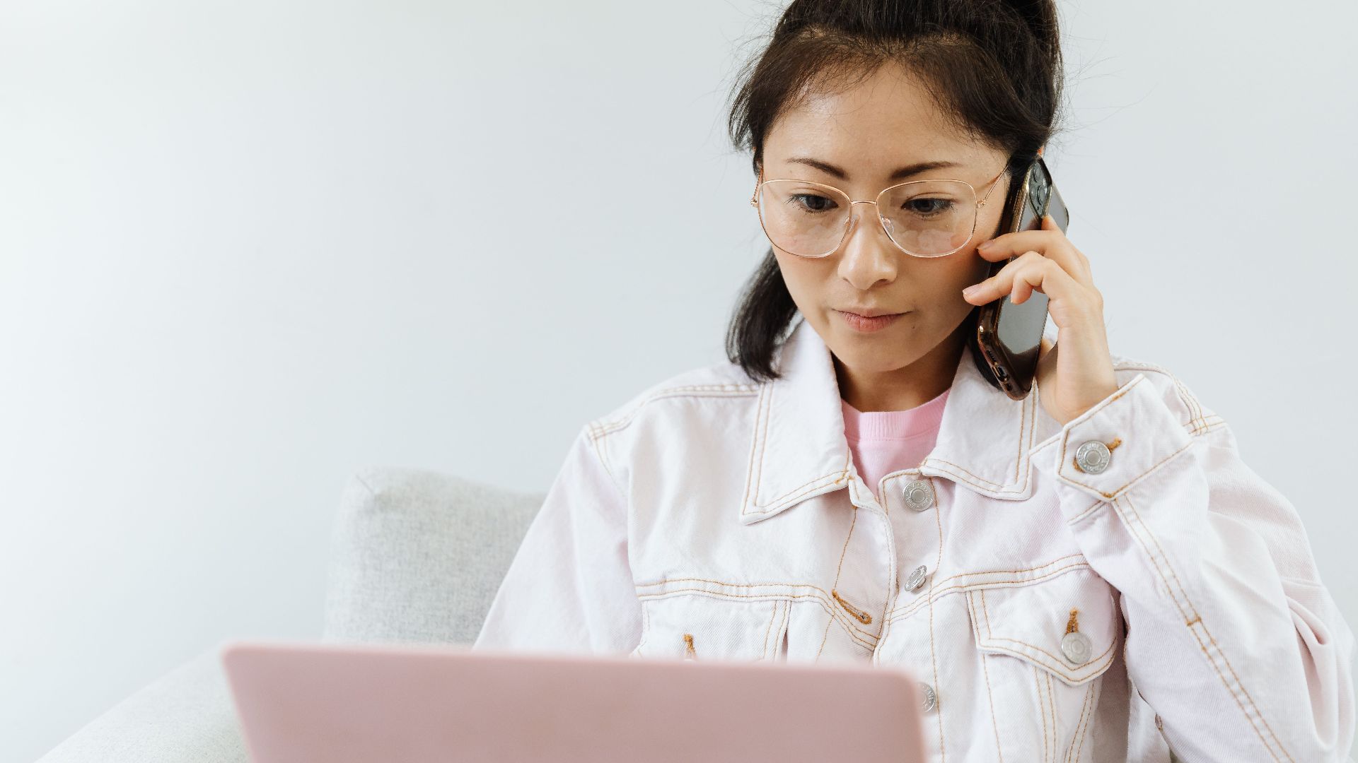 Woman in glasses multitasking with phone and laptop in a serene office setting.