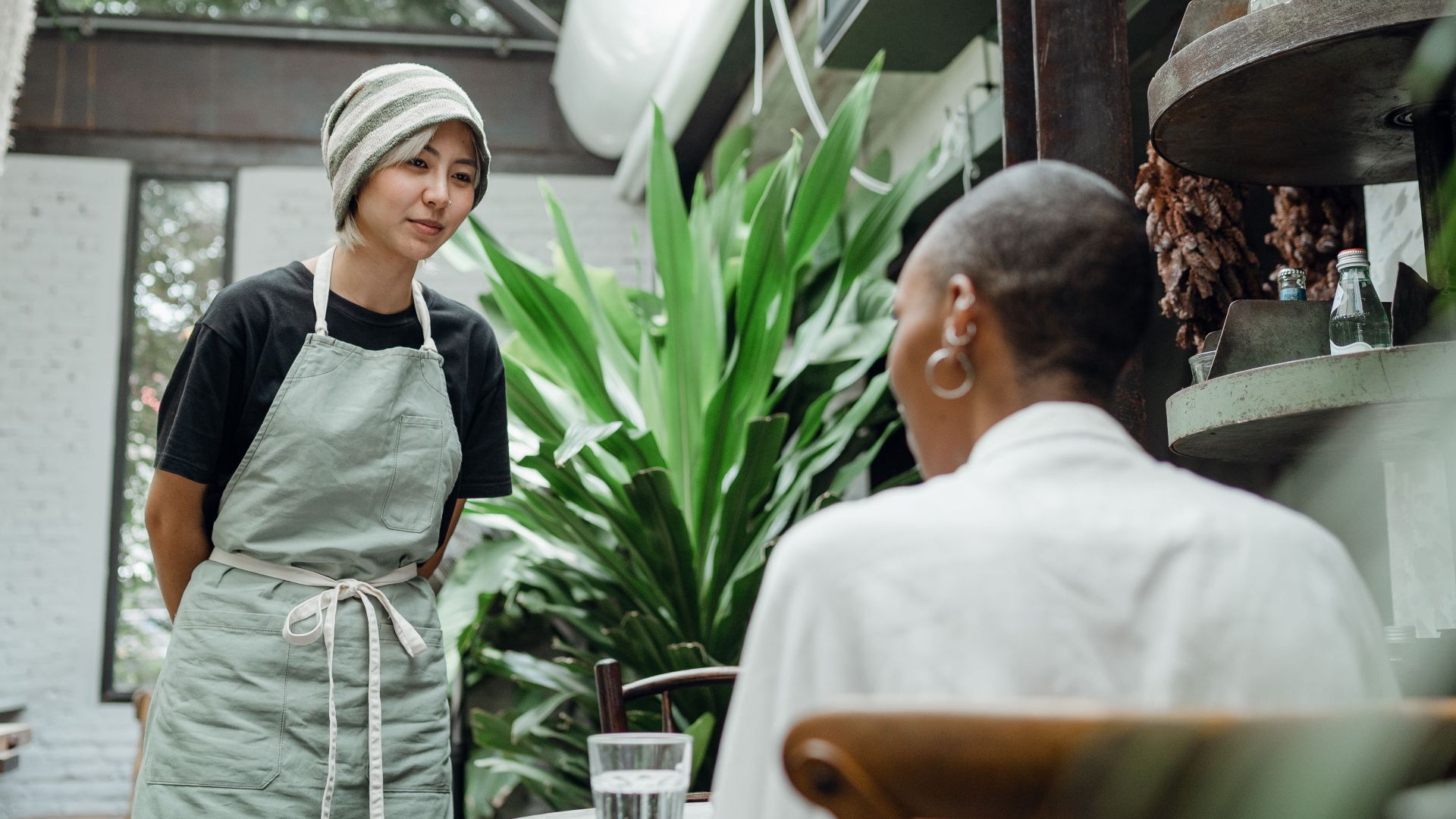 Friendly young Asian waitress in apron and hat standing near table and talking to female customer while working in cozy cafe