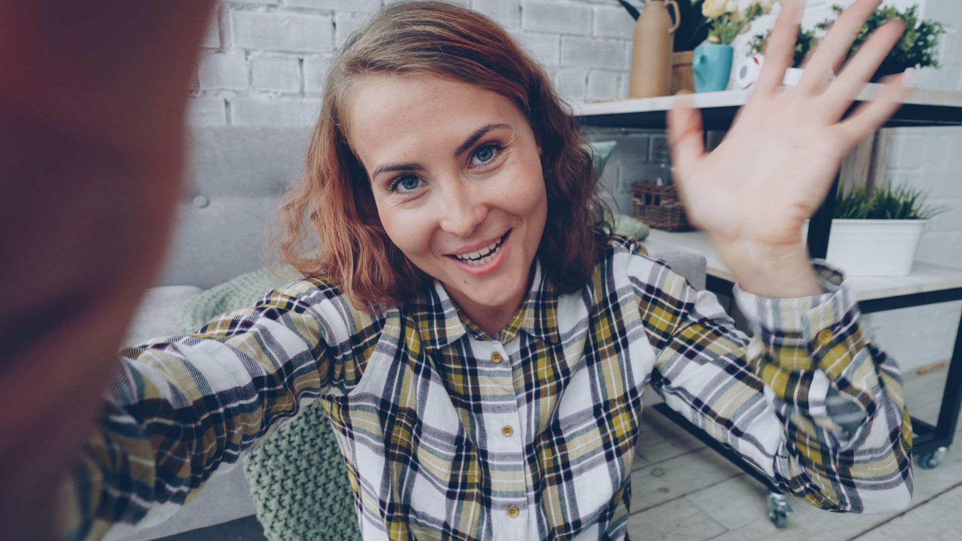 Cheerful woman in plaid shirt waving in a cozy indoor setting. Perfect for lifestyle or social media themes.