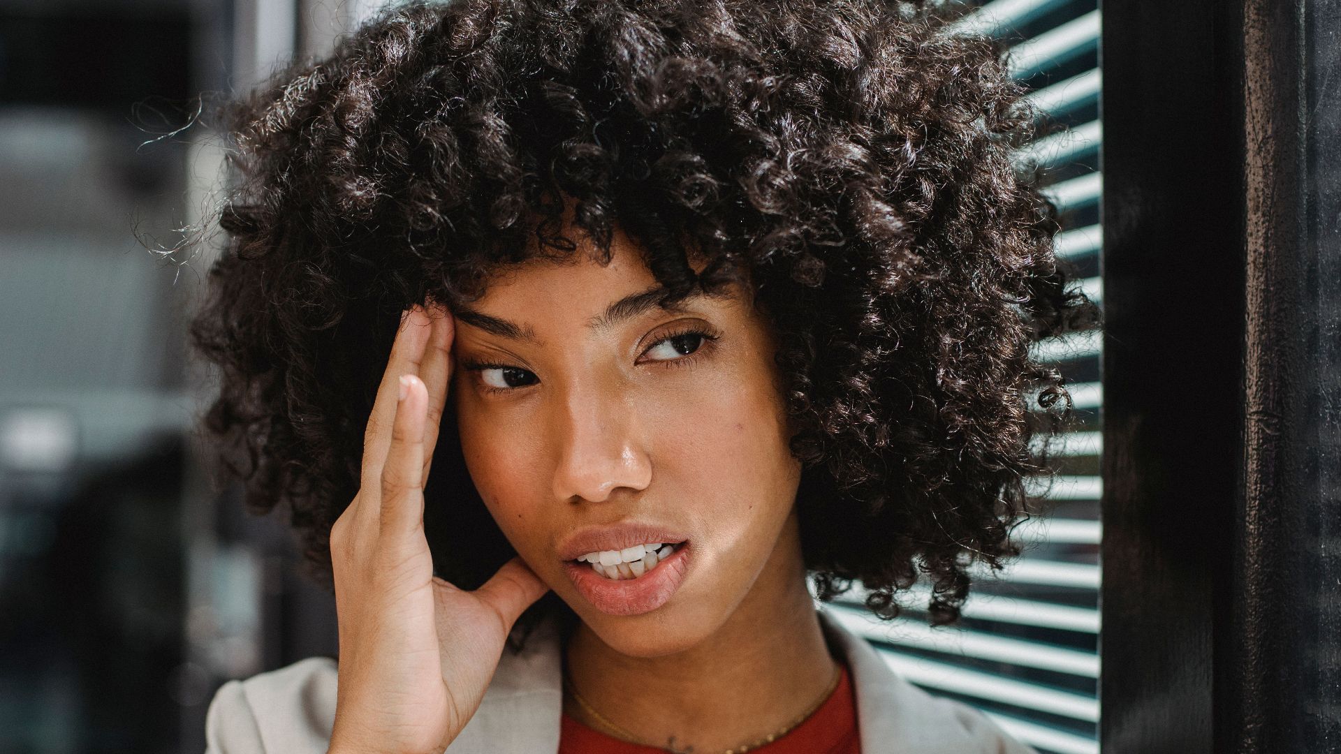 A businesswoman with curly hair expresses stress by her office window, touching her forehead.