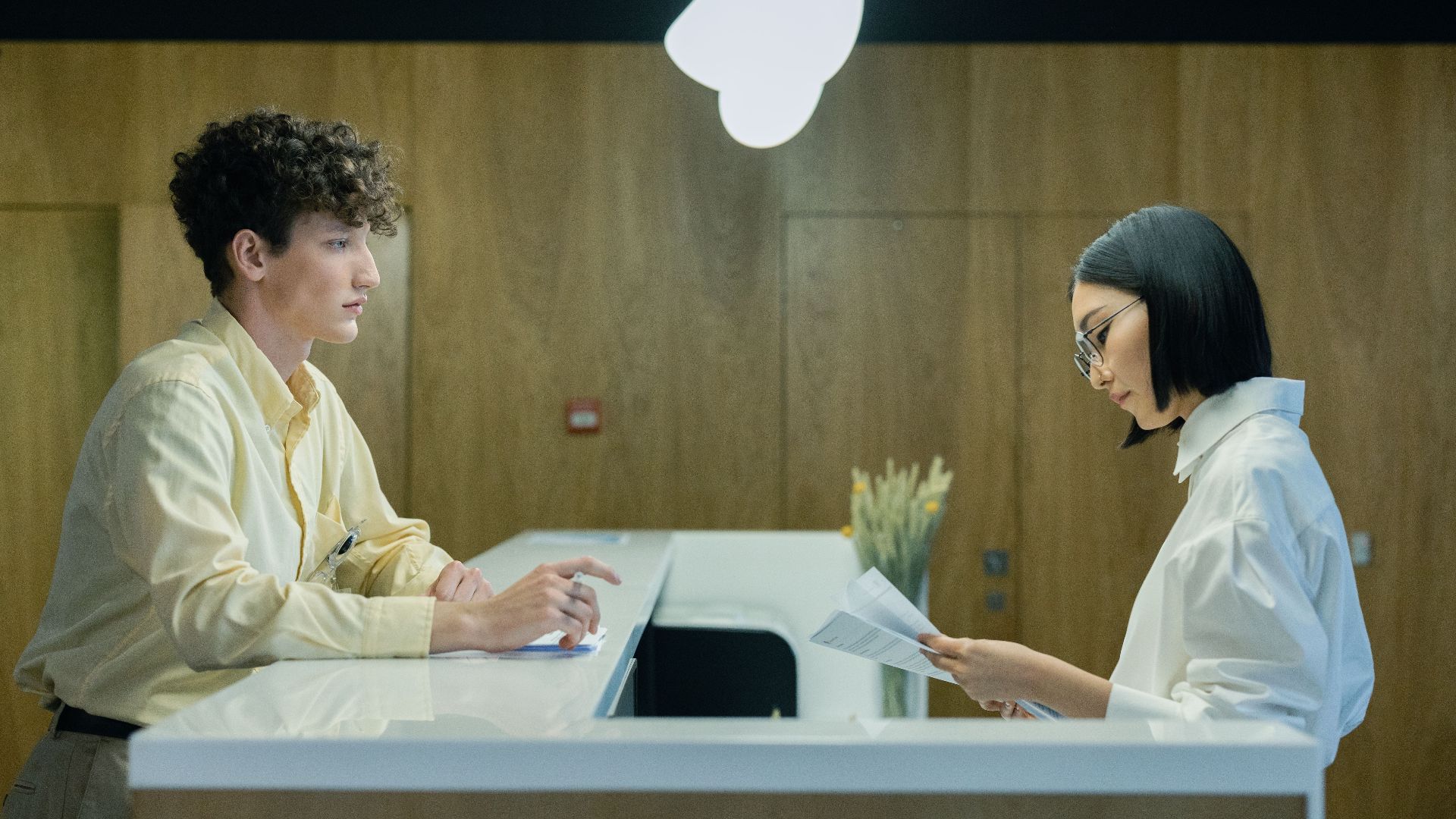 A young man and woman interacting at a modern office reception desk indoors.