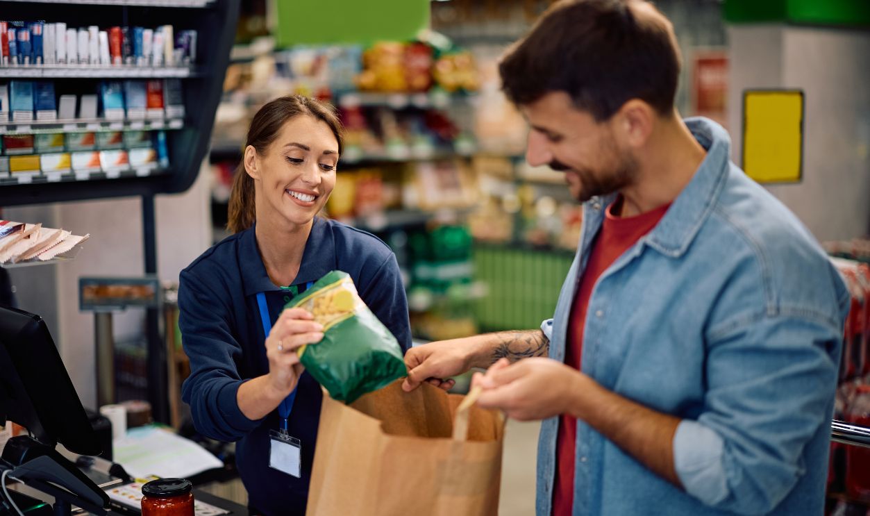 Shutterstock - 2645728023 - Happy supermarket cashier assisting a customer in packing groceries in paper bag at the checkout.