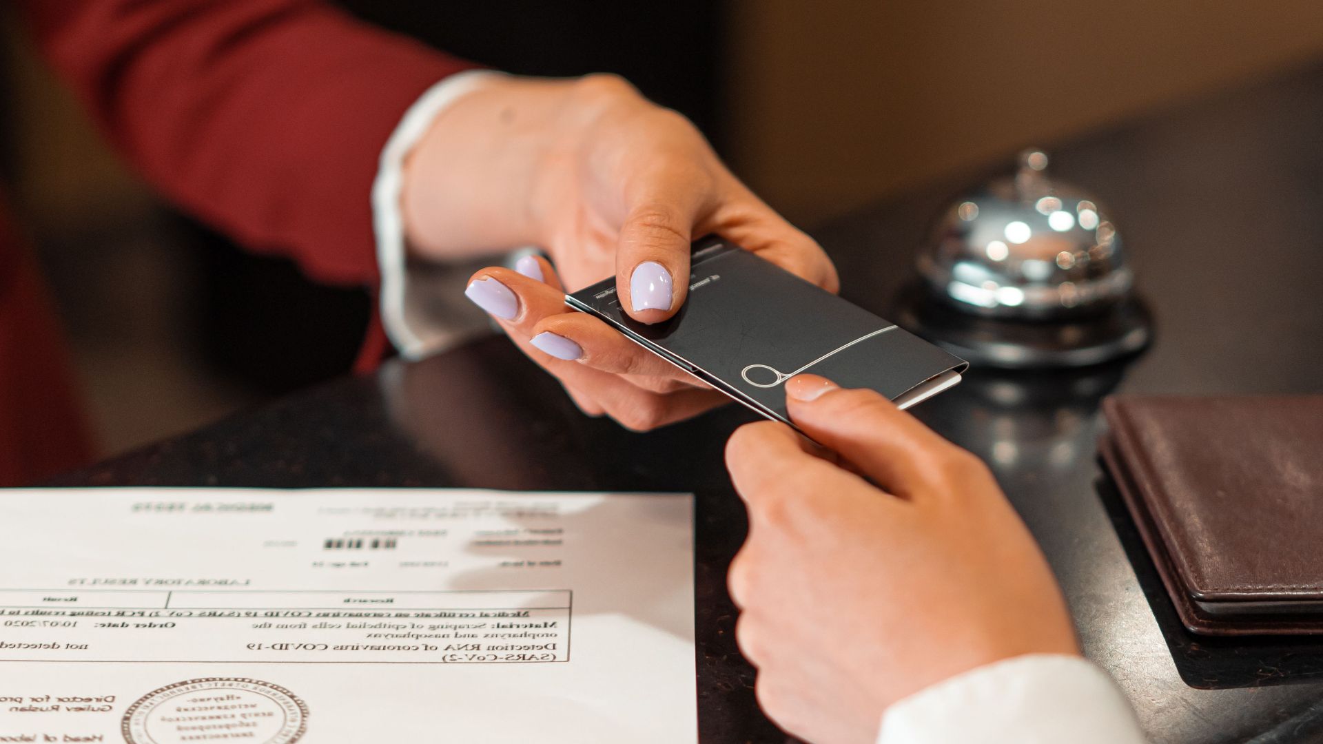 A hotel receptionist hands a key card to a guest over the counter, with documents visible.