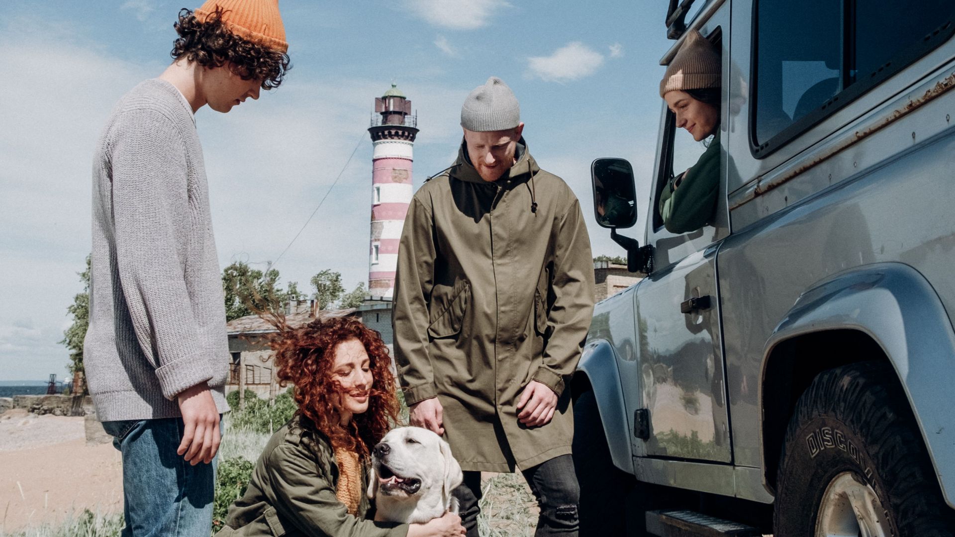 Friends and a dog relax beside a car near a lighthouse on a sunny beach day.