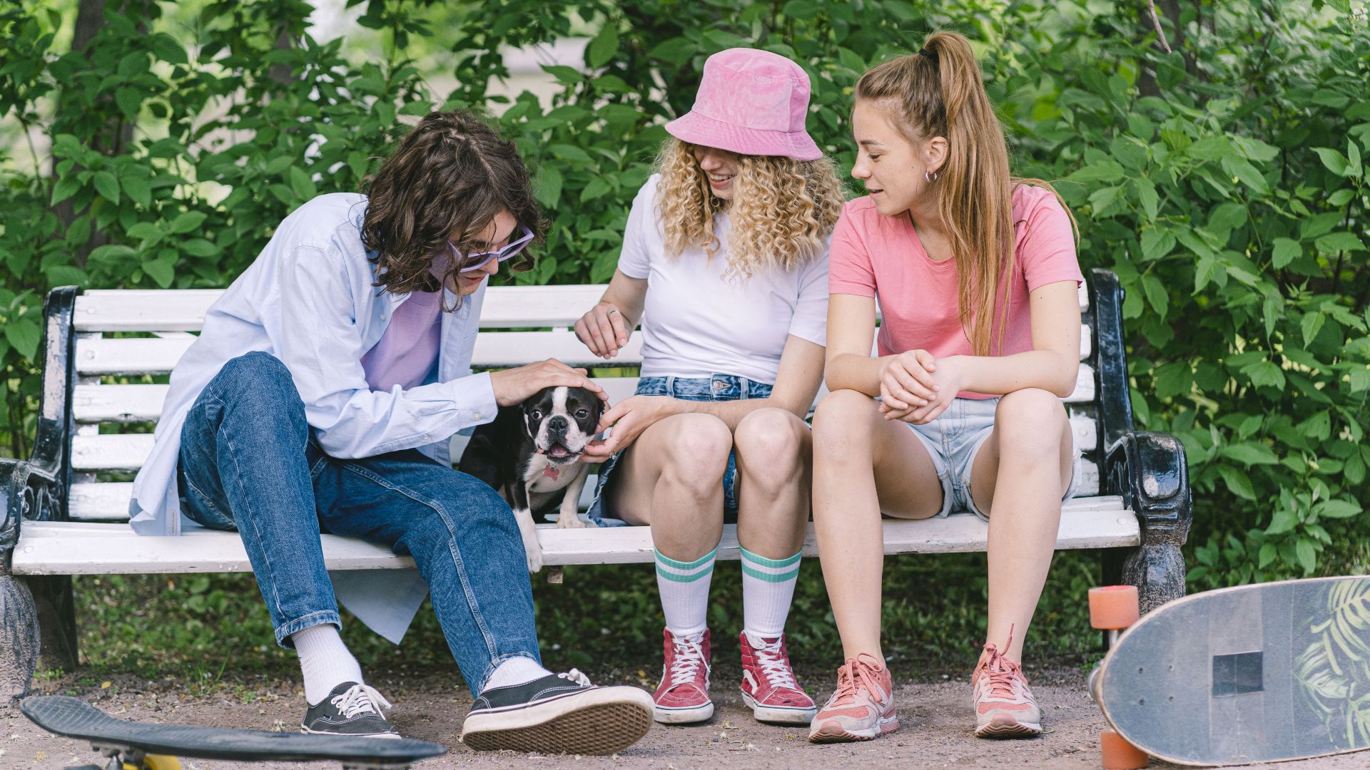 Three friends enjoy a moment with a dog on a park bench, accompanied by skateboards on a sunny day.