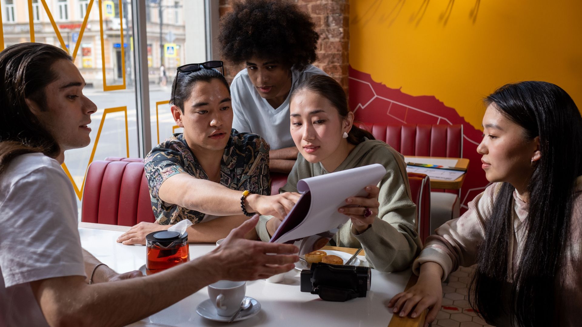 A multicultural group of young adults engaged in conversation around a table in a cafe, focused on a document.