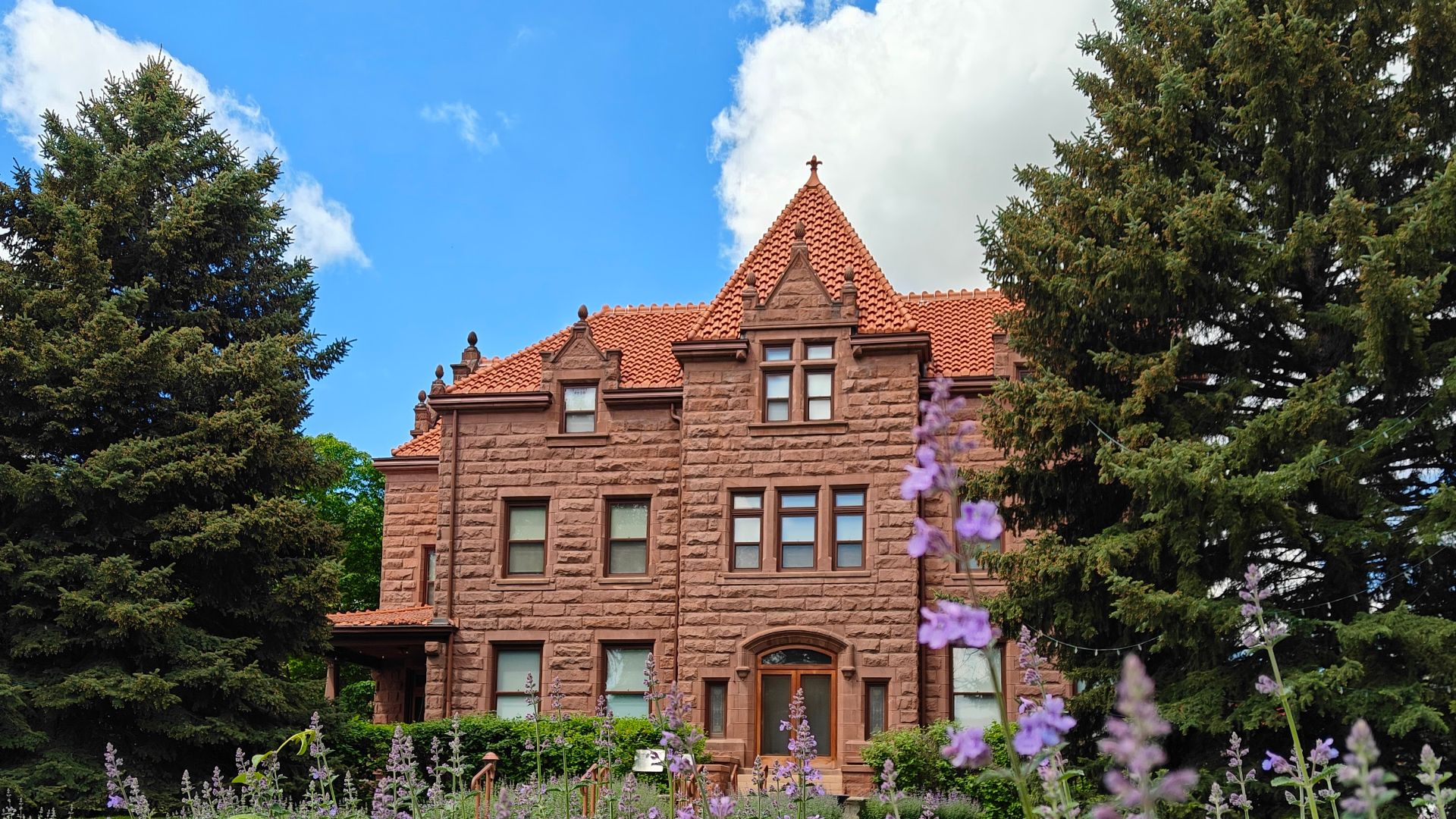 The front of the Moss Mansion in Billings, Montana, as seen through the sidewalk hedges. The flowers are some variety of catmint.