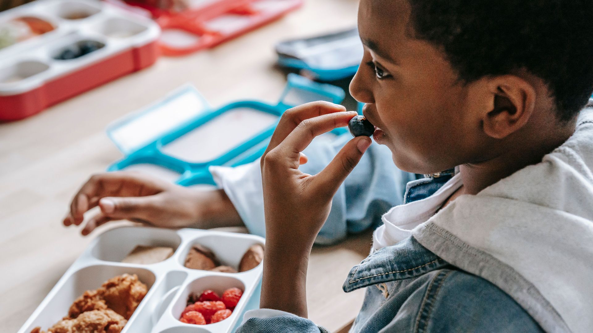 Crop African American child in casual clothes sitting at table with plastic lunch box while having snack