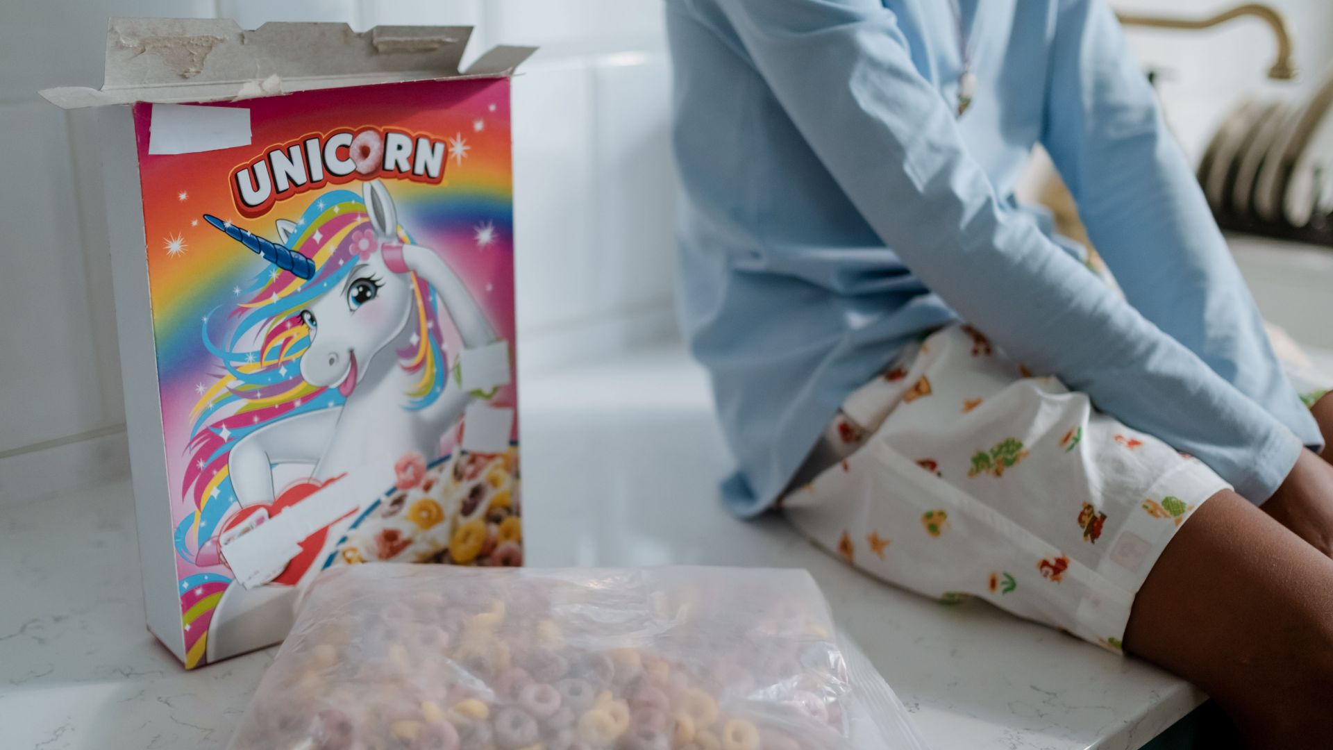 A child sitting on a kitchen counter with an open box of unicorn-themed cereal, highlighting a playful morning scene.