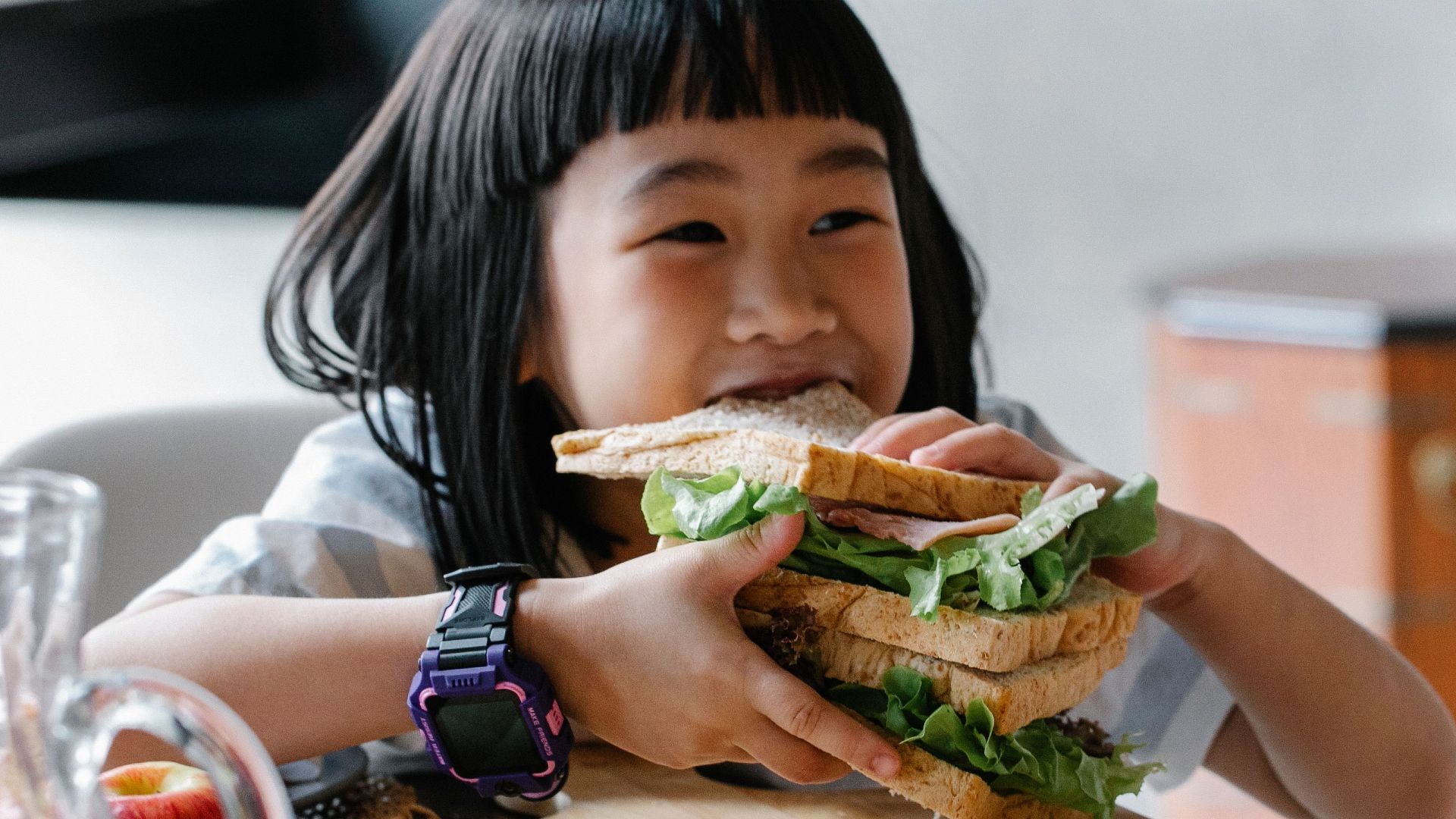 Asian child sitting at wooden table during breakfast and eating tasty sandwich with lettuce leaves
