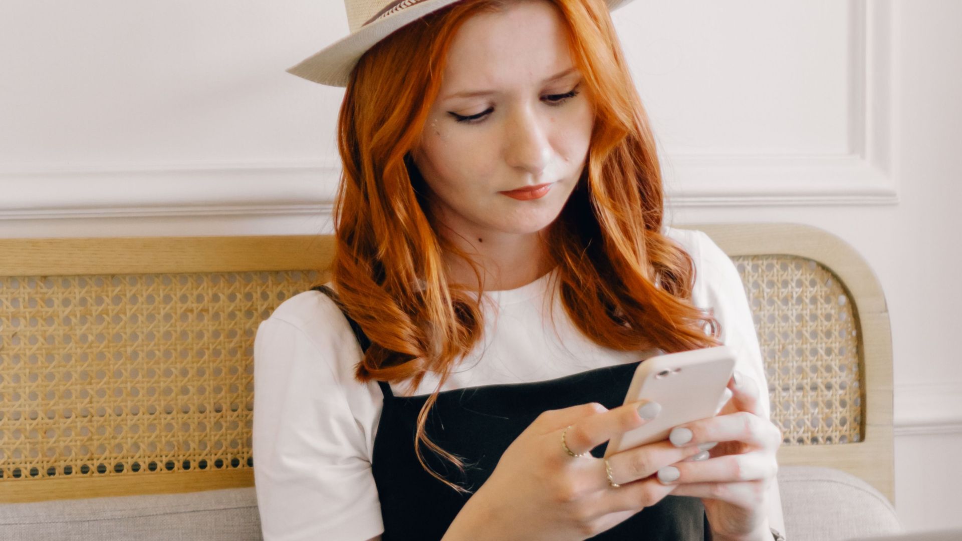 A young woman in a fedora hat texting on her smartphone while sitting with a laptop indoors.
