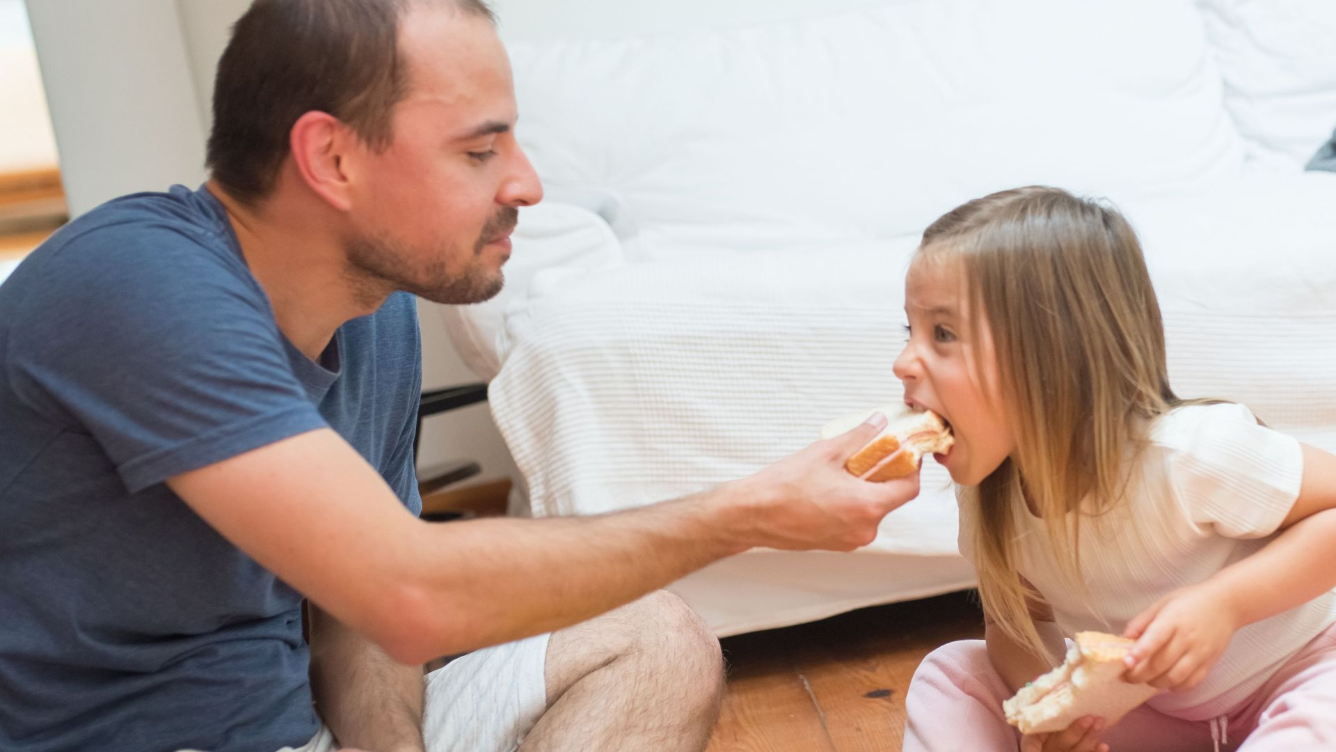 Father and daughter bonding over a snack on a wooden floor. Warm family moment captured indoors.