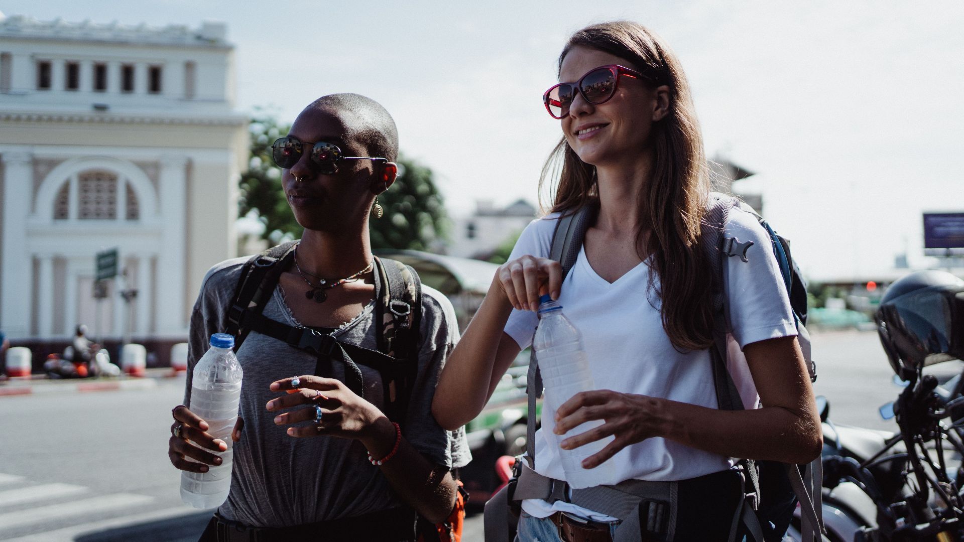 Two women travelers enjoying a sunny day, holding water bottles, wearing sunglasses and backpacks.