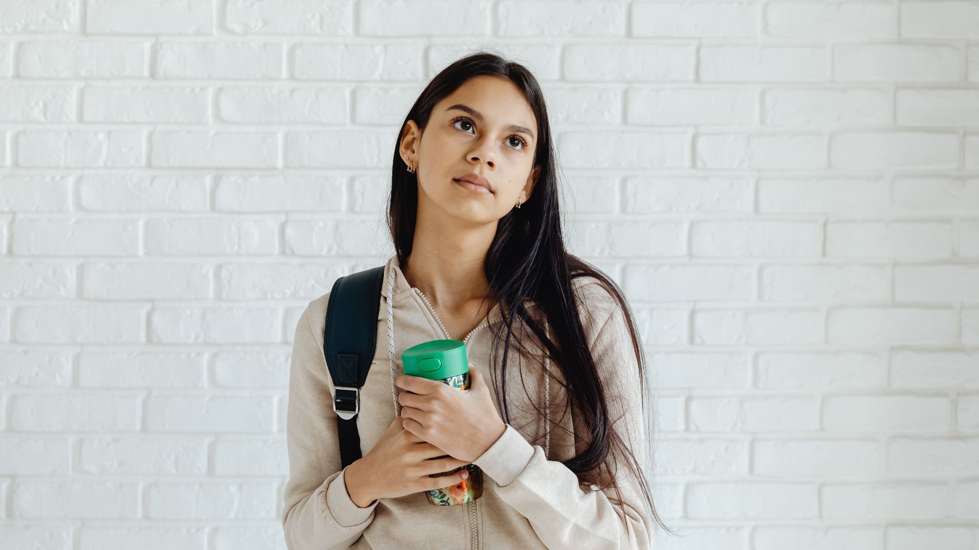 A pensive young woman in a hoodie, holding a tumbler, leans on a white brick wall.