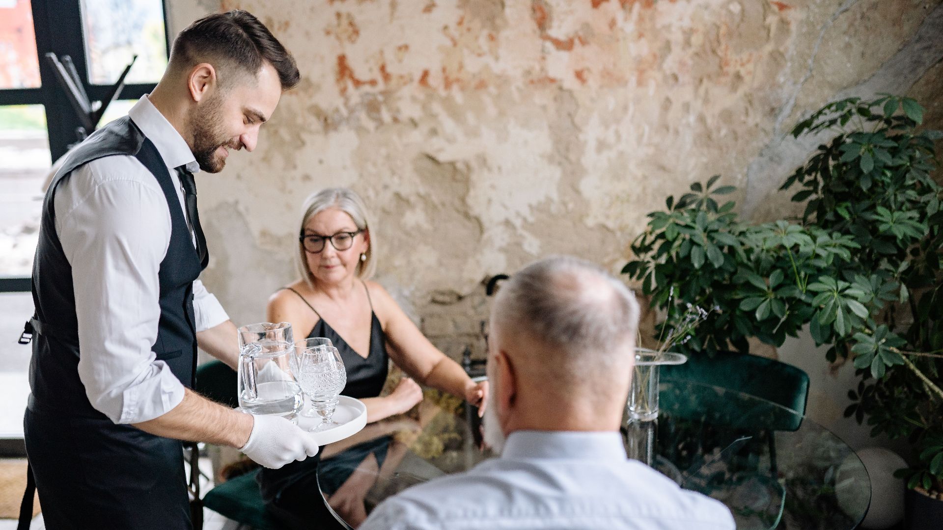 A waiter serving drinks to a senior couple in a stylish, rustic restaurant setting.