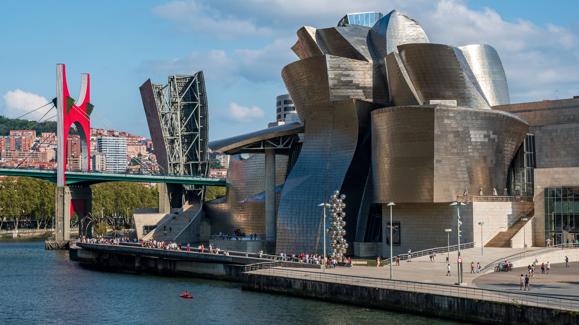 Guggenheim Museum Bilbao from Northwest. Three sculptures from the museum's collection can be seen outside: Tulips by Jeff Koons, Tall Tree & The Eye by Anish Kapoor, and Maman by Louise Bourgeois. The Salve Bridge stands out with its red portico.