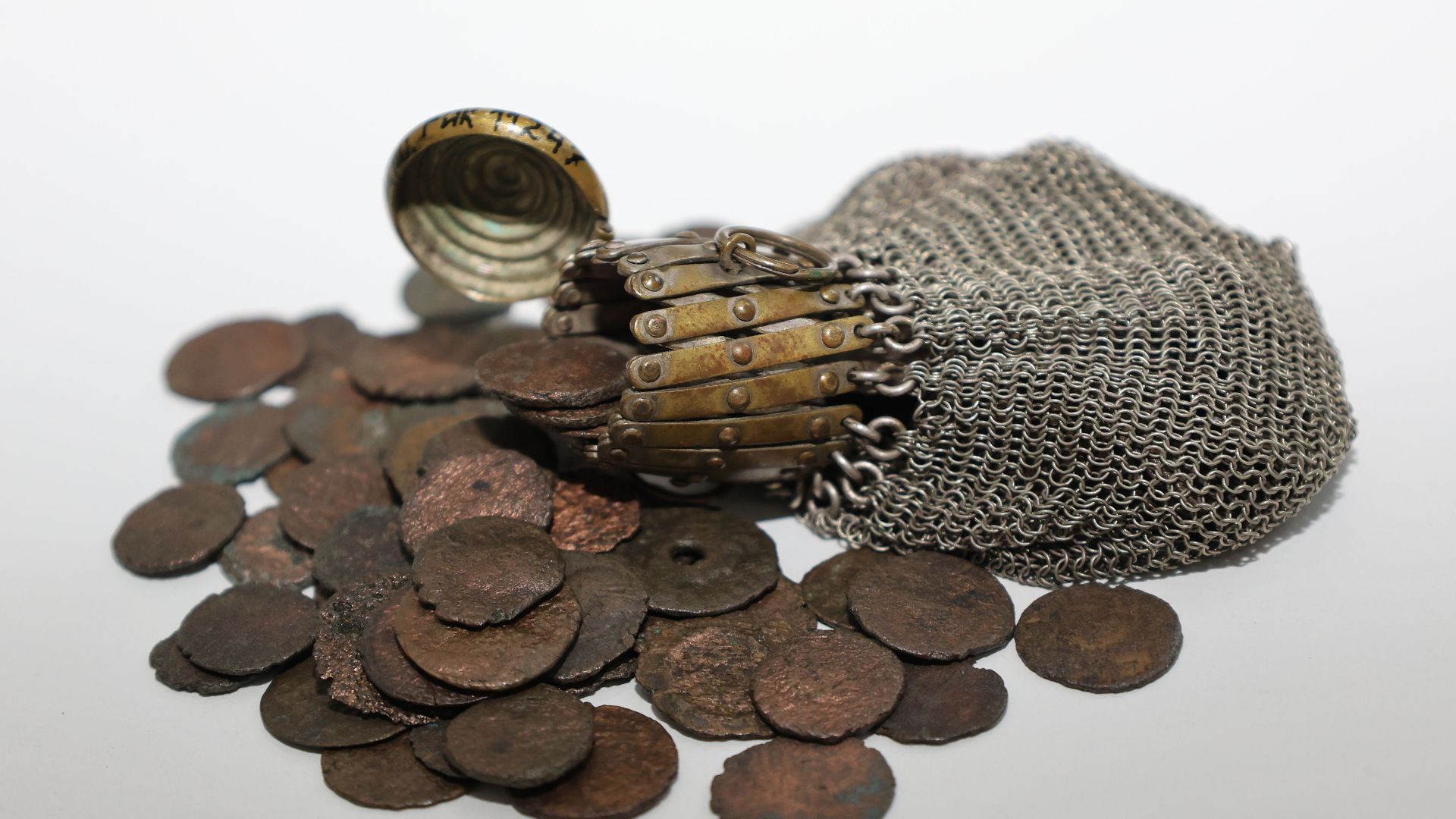 Antique metal chain pouch spilling ancient rustic coins on a white surface.