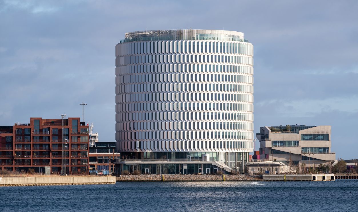 Copenhagen, Denmark - March 1, 2026: The award-winning Tip of Nordo cylindrical office tower rises above the Nordhavn waterfront on a clear winter morning, its 925 faceted glass and galvanized steel facade elements catching the light to create a shimmering textured surface visible from across the harbor. Designed by Cobe, Vilhelm Lauritzen Architects and Third Nature, the 60-meter-tall silo-shaped landmark was completed in 2023 and won the Copenhagen Building Award 2025. The building houses law firm Bech-Bruun and Copenhagen Infrastructure Partners across 25,000 square meters of flexible office space. Flanking the tower, contrasting architectural styles illustrate Nordhavn's diverse urban development: a red brick Residence Inn hotel on the left echoes traditional warehouse design, while a brutalist concrete office building on the right adds modernist contrast. The cylindrical form references the historic grain silos that once defined this stretch of Copenhagen's industrial waterfront along the Sound. Nordhavn is one of Europe's largest urban regeneration projects, transforming a former container port into a mixed-use district planned for 40,000 residents and 40,000 jobs. The clear winter light and calm harbor water provide an unobstructed view of the facade's distinctive angled louver system, designed to regulate solar heat gain while creating the building's signature visual rhythm. Suitable for architecture editorial, urban planning features, Scandinavian design publications, and Copenhagen travel content.