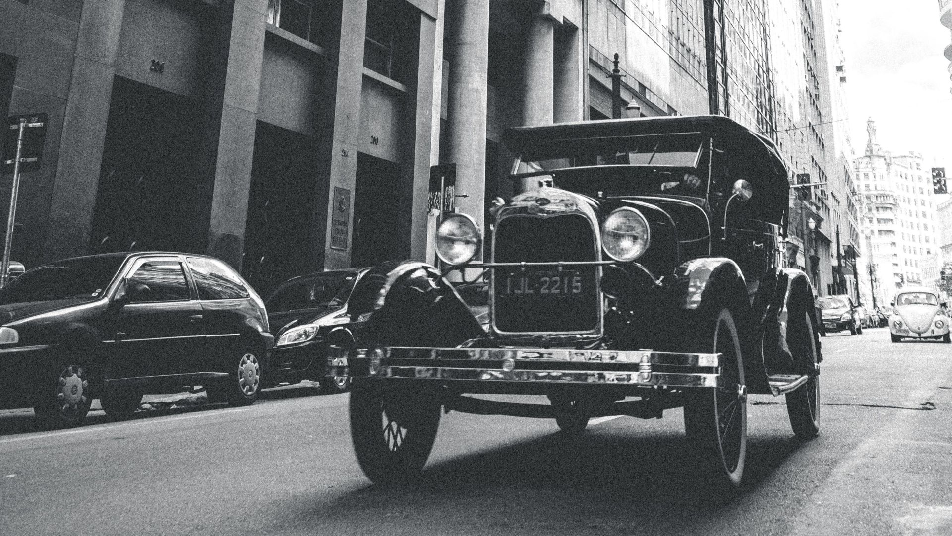 A vintage car drives through an urban street lined with tall buildings, under a cloudy sky.