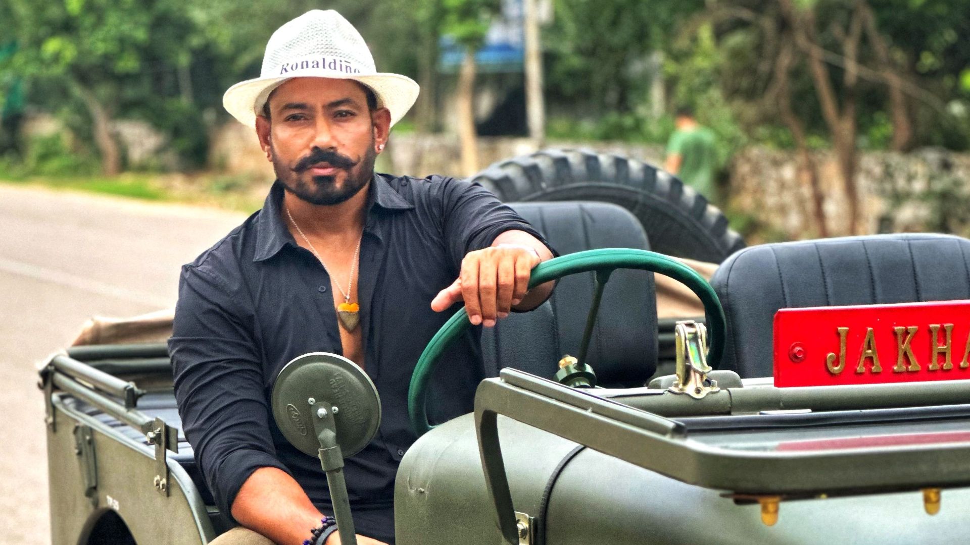 A man in a vintage jeep wearing a fedora enjoys an off-road adventure on a rural road.