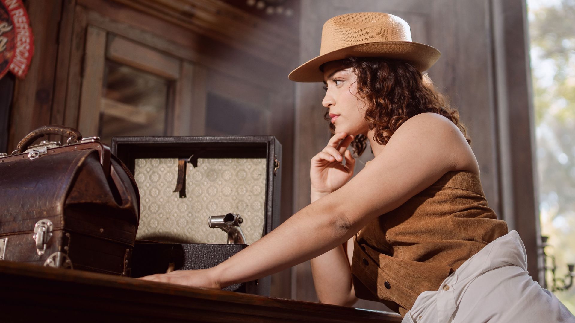 A woman in vintage attire poses thoughtfully in a sunlit room filled with nostalgic decor.