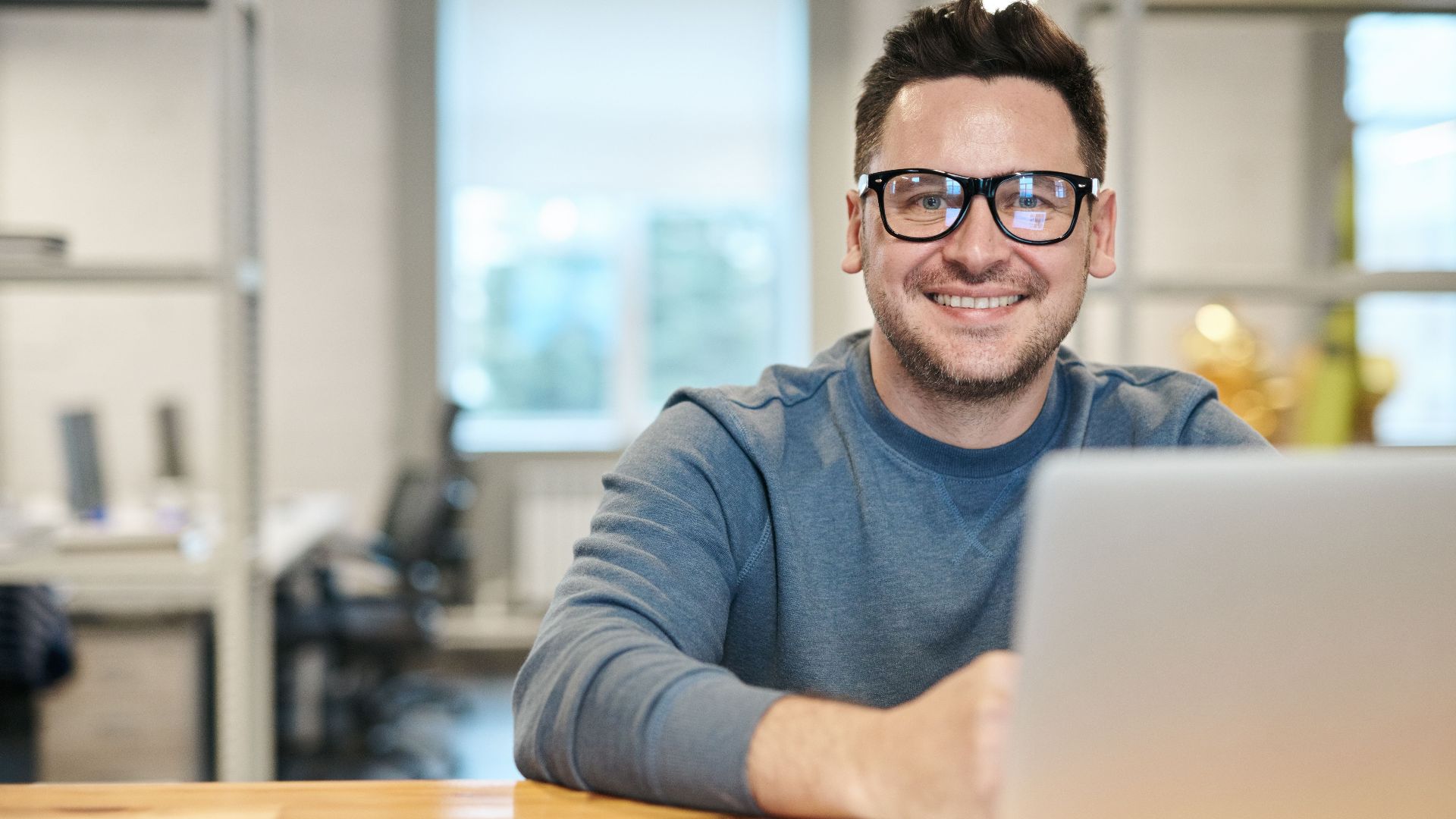 Happy man wearing glasses working remotely on laptop in modern office environment.