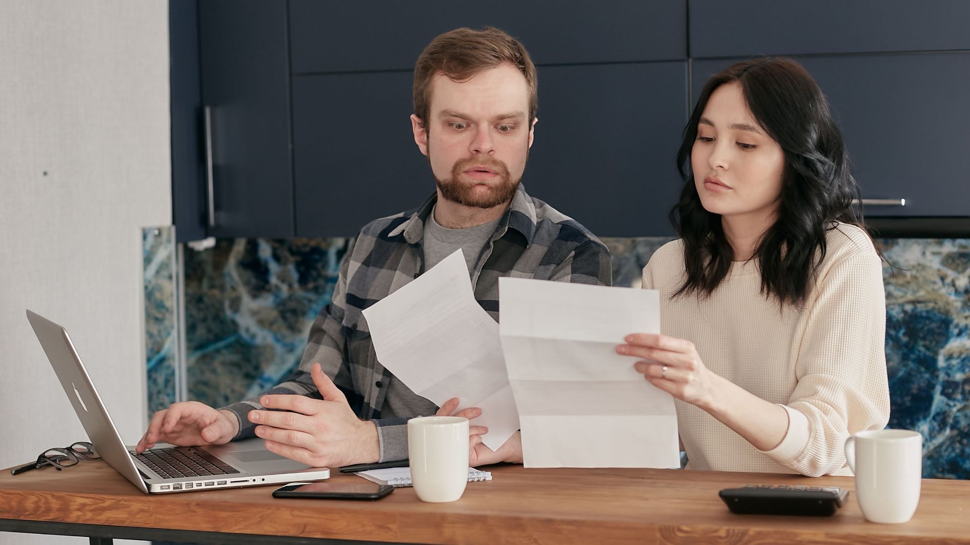 A couple sitting at a wooden table, reviewing documents with a laptop nearby, expressing concern.