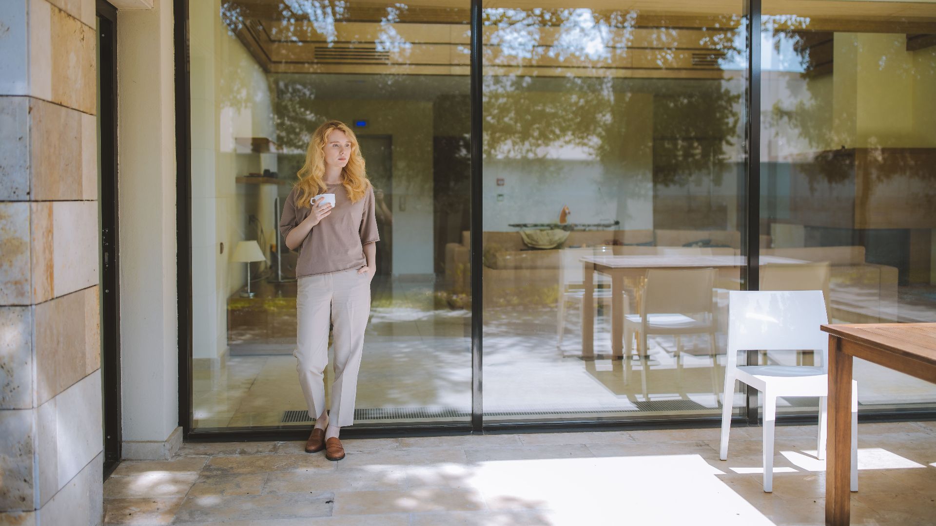 Stylish woman enjoying a coffee while standing by a glass wall in a modern, sunlit room.