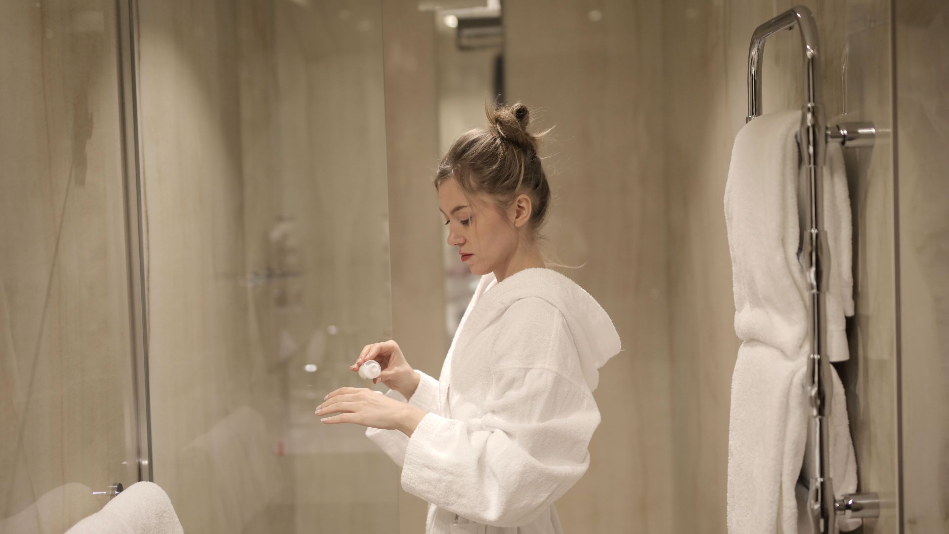 A woman in a bathrobe applies lotion in a contemporary bathroom with glass doors and towels.