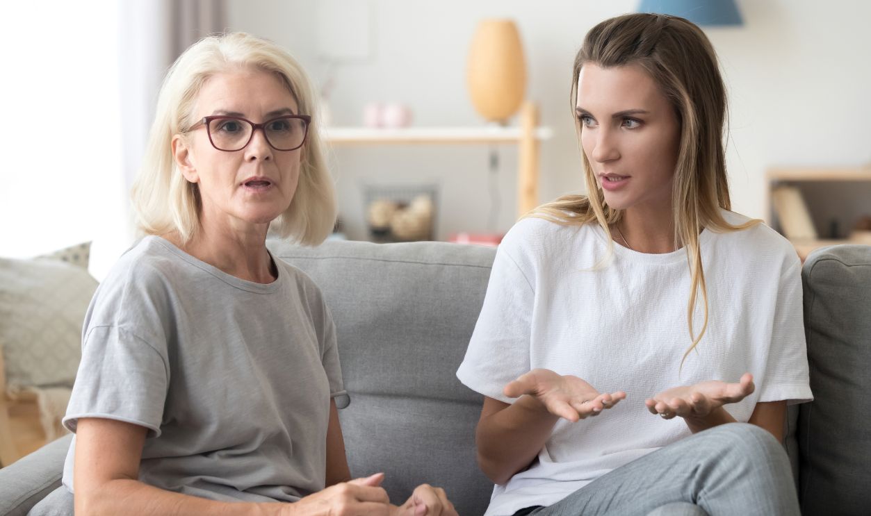 Middle aged woman sitting on couch in living room with young female at home. Serious mother and young daughter family people discussing arguing talking together indoors having unpleasant conversation