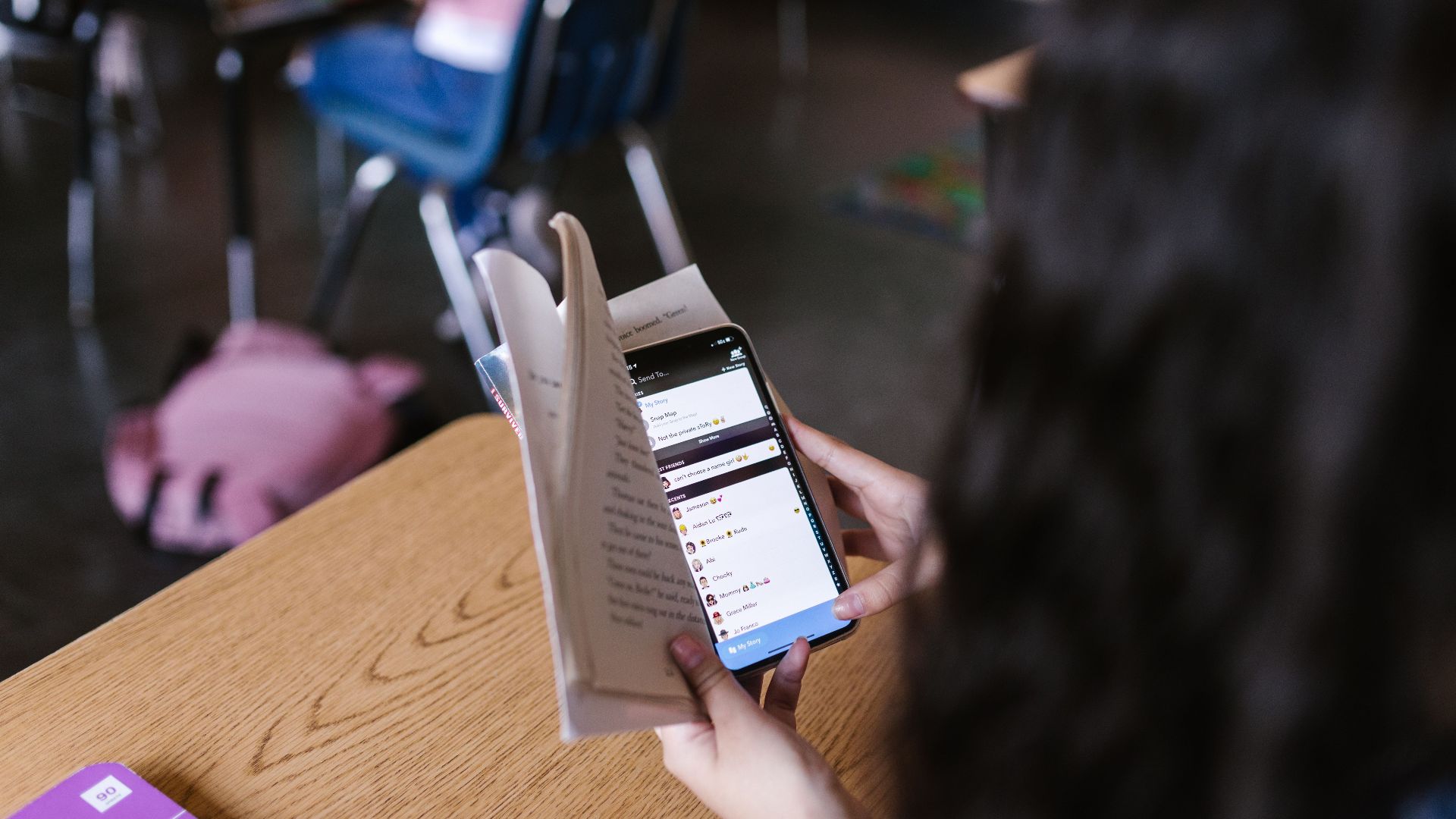 A student in a classroom multitasks by reading a book while using a smartphone, emphasizing modern educational challenges.
