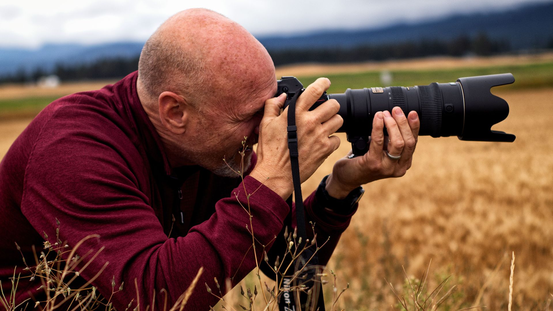 A bald photographer takes nature shots in a field with a DSLR camera.